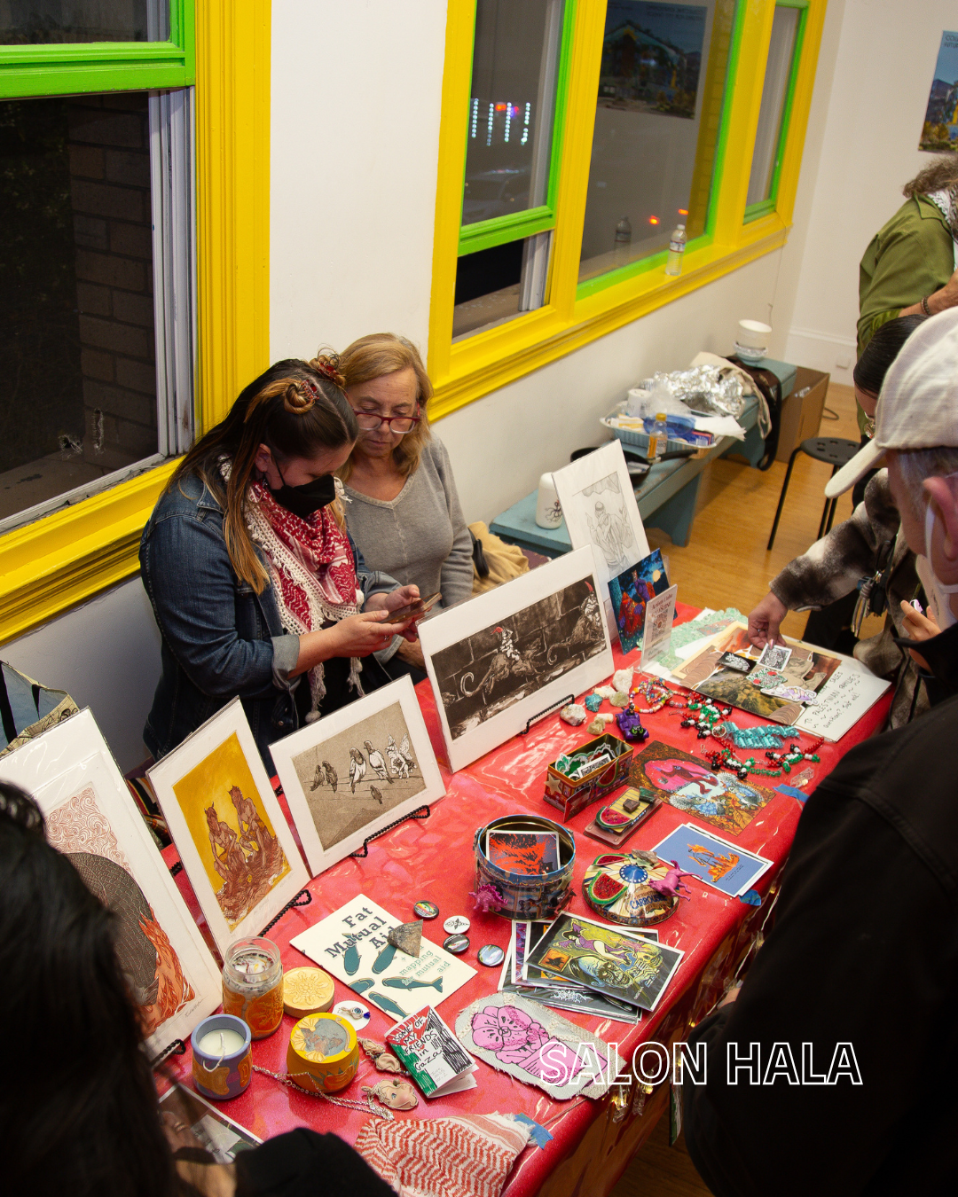 Art and craft booth at an indoor market with artwork, jewelry, and small items on a red tablecloth, with people browsing and talking.