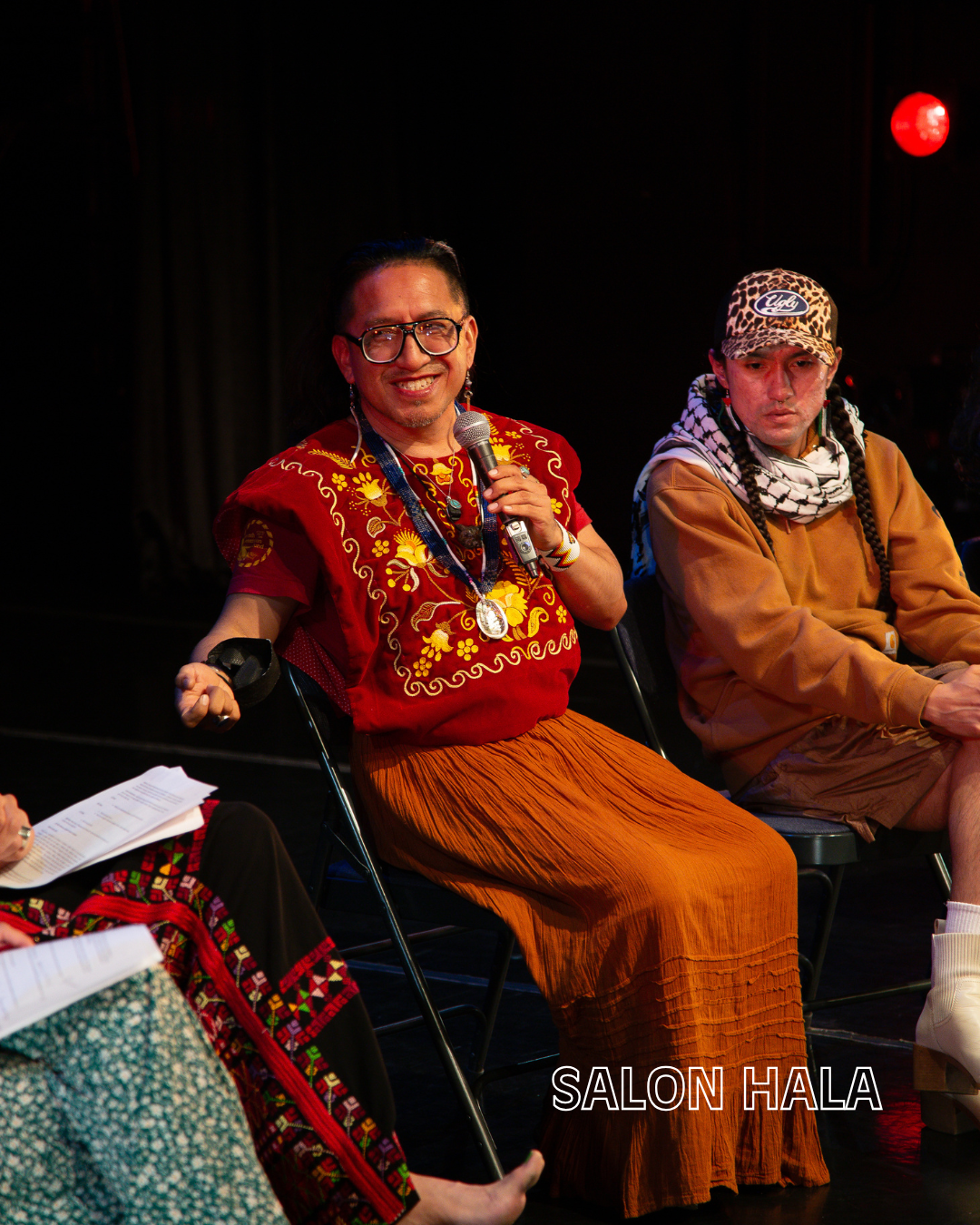 A woman in colorful traditional clothing holding a microphone, smiling, seated next to a man in casual clothing with braids, wearing a leopard-print cap, on a stage with a black curtain background and red lighting. The text "SALON HALA" is at the bot