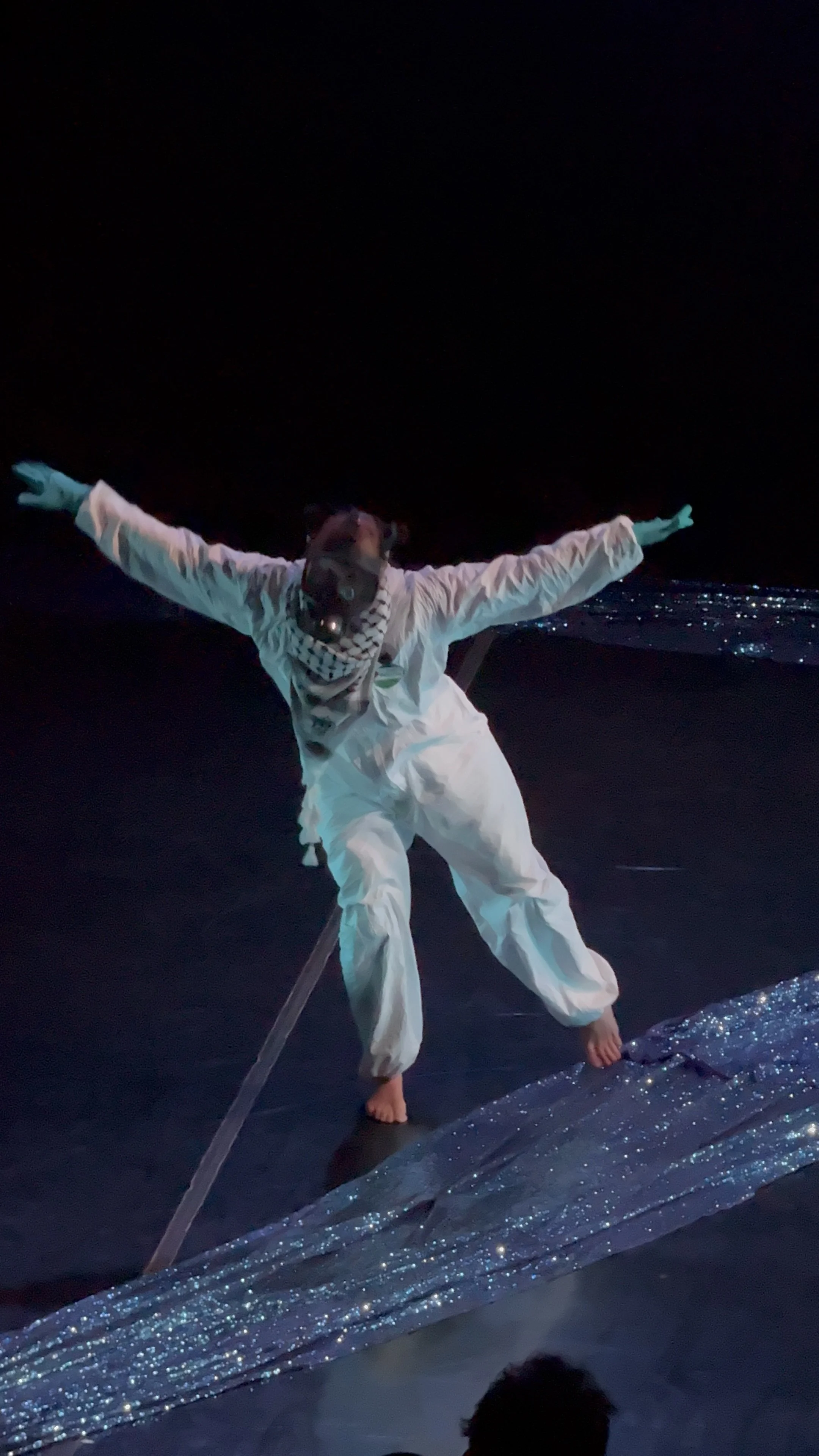 Person dressed in white standing on a glittery surface at night, with arms outstretched and a dark background.