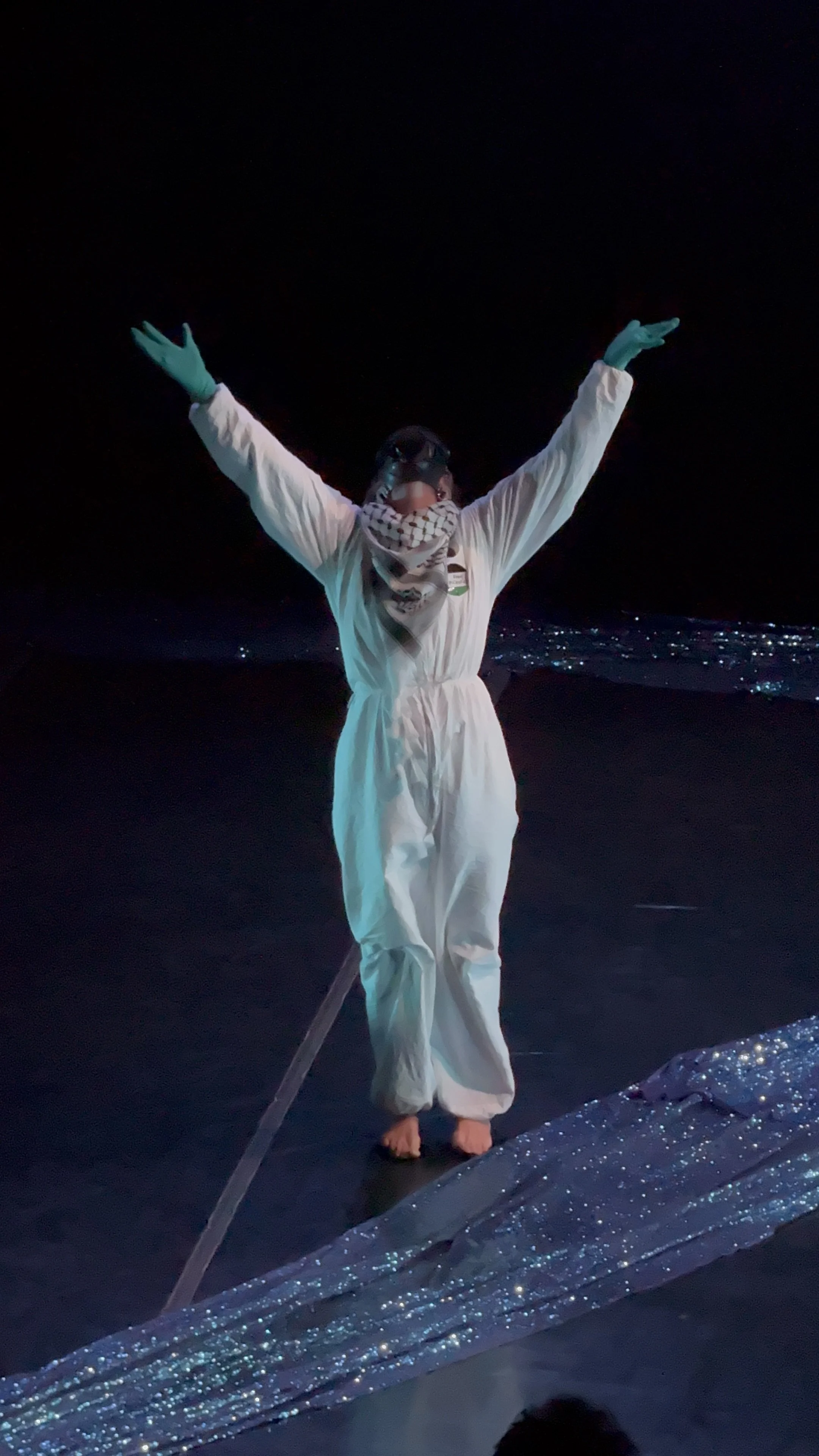 Person dressed in white protective suit, face mask, goggles, and gloves, standing in the dark with arms raised, on a glittery surface near water.