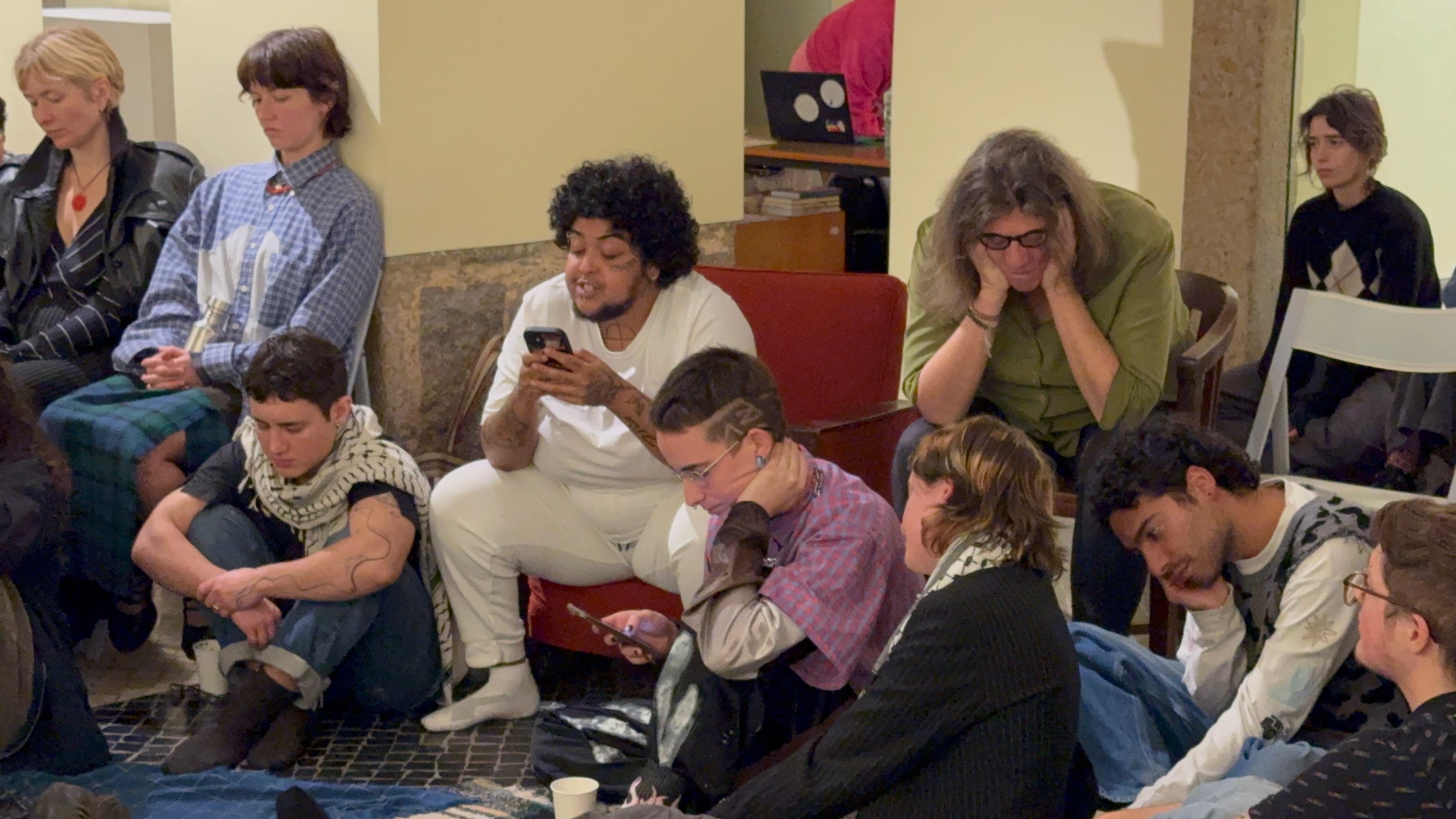 People sitting on the floor and chairs in a room, some looking at their phones, others with eyes closed or head resting on hands, appearing tired or bored.