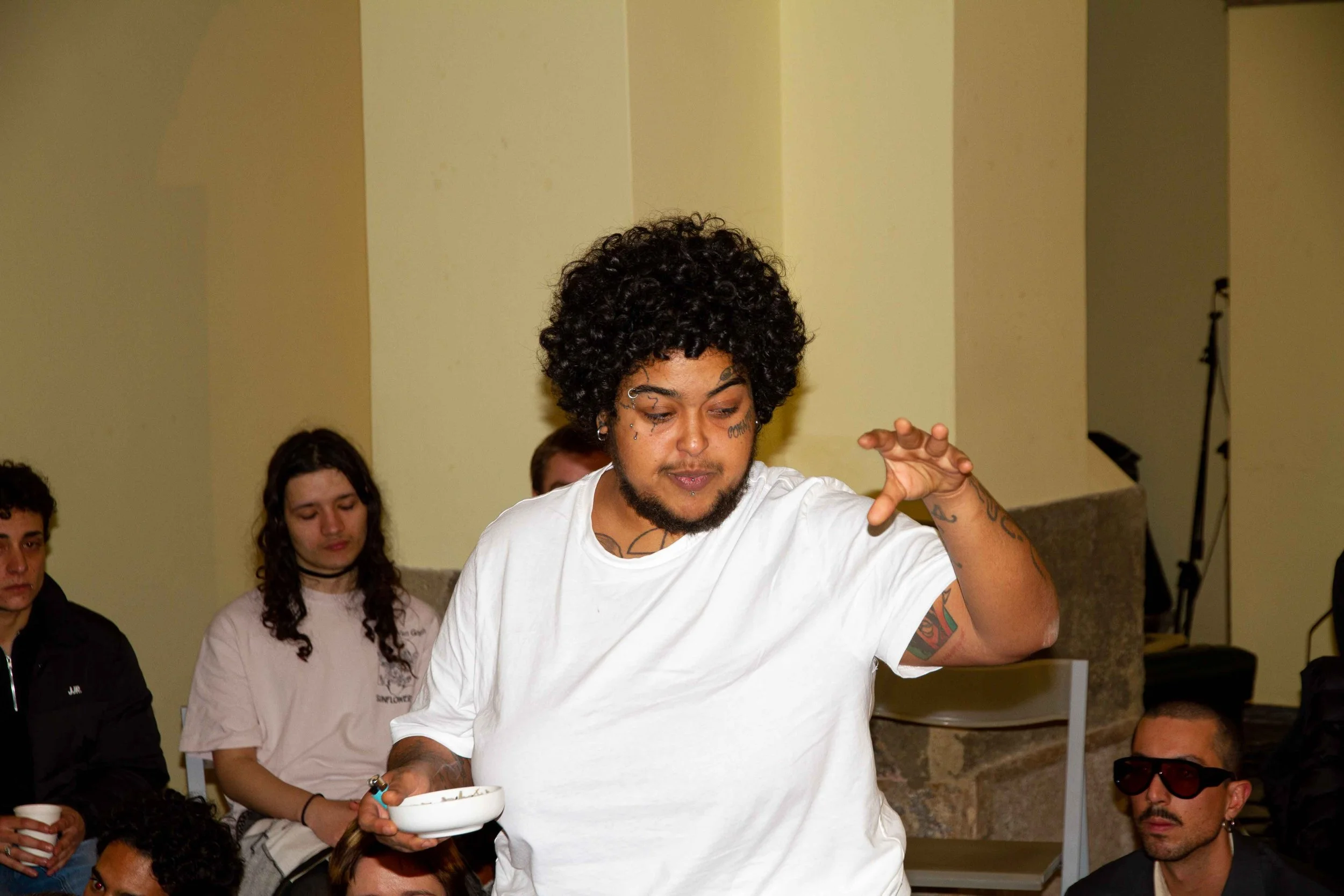 A man with curly hair and tattoos on his arms is holding a bowl and appears to be speaking or gesturing during a group meeting or discussion, with several people seated around him.