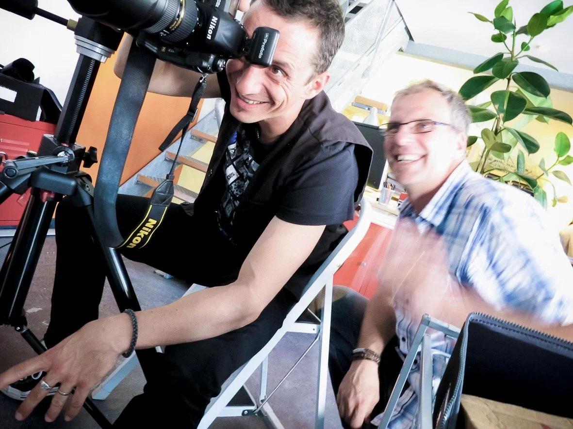 Deux hommes souriants, l'un utilisant un appareil photo Nikon sur un trépied, dans un environnement intérieur avec une plante verte.