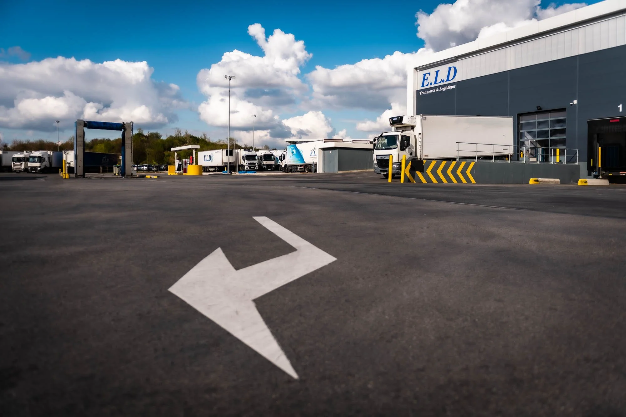 Parking avec flèche directionnelle et camions, bâtiment industriel à l'arrière-plan.