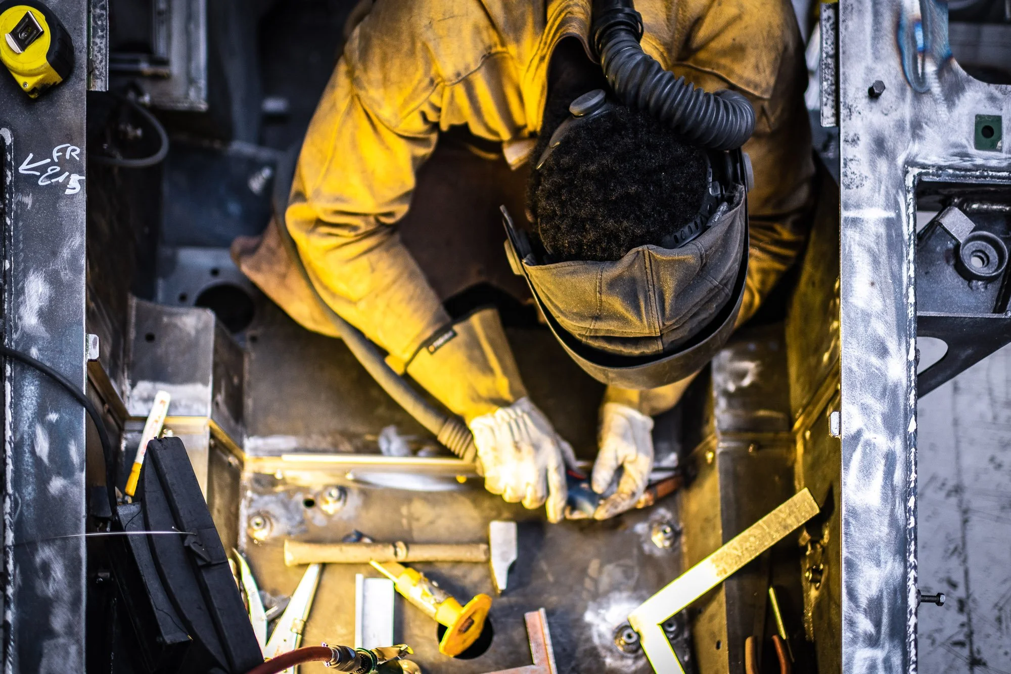 Une personne en tenue de protection effectue une tâche de soudage, entourée d'outils métalliques, dans un environnement industriel.