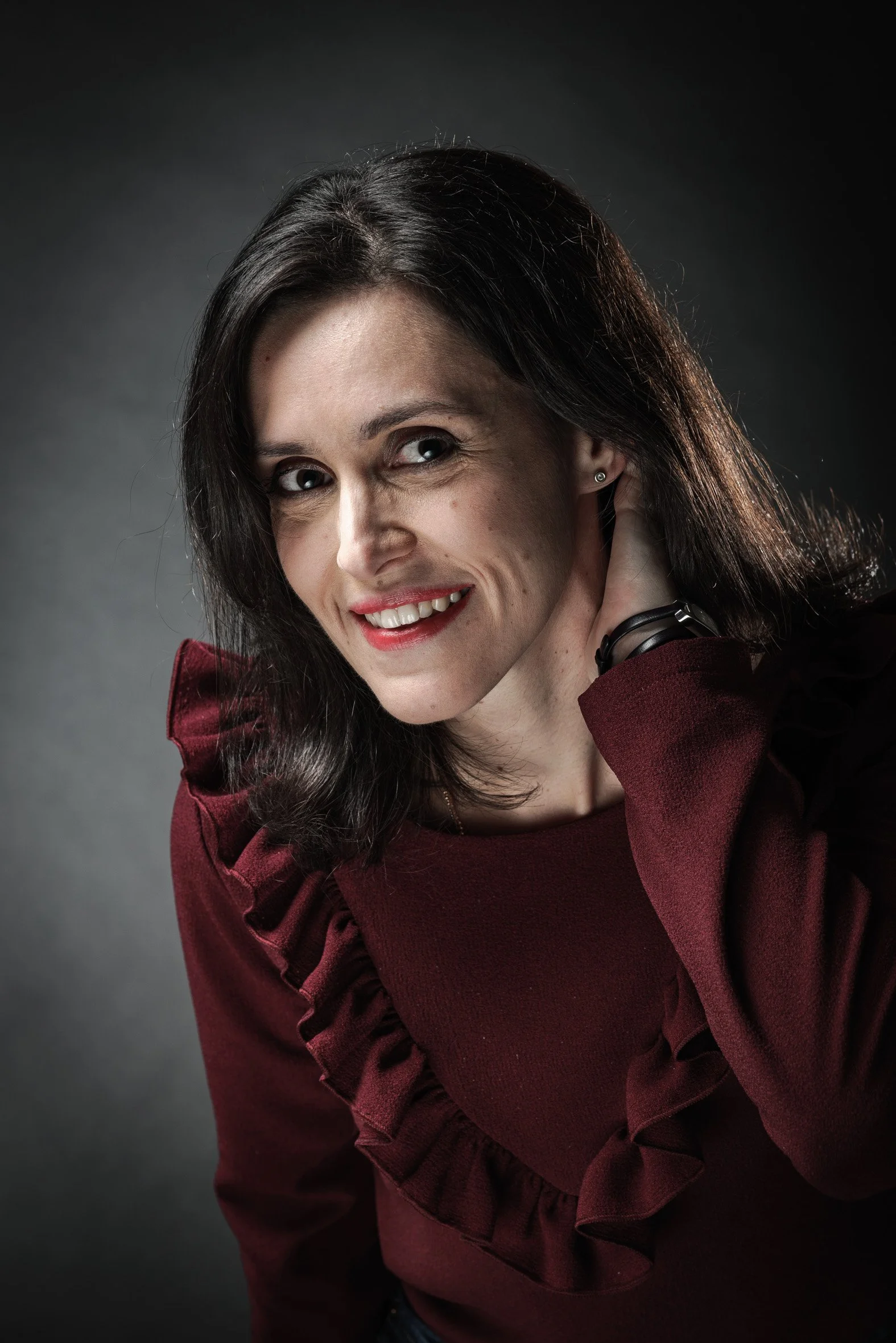 Femme souriante, cheveux noirs, portant un pull rouge bordeaux, pose devant un fond sombre.