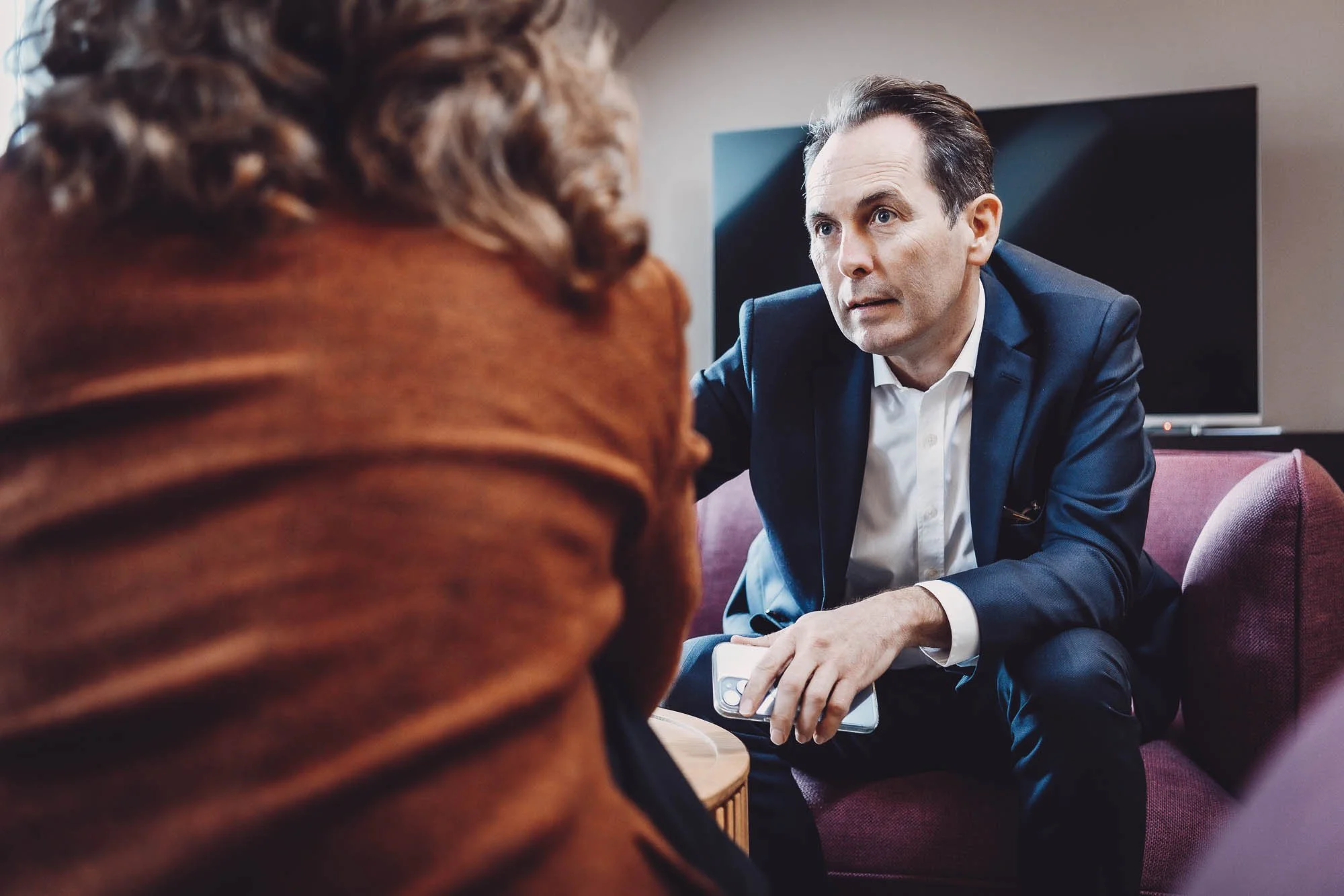 Un homme en costume discute avec une personne en veste marron, assis dans un salon moderne avec un téléviseur en arrière-plan.