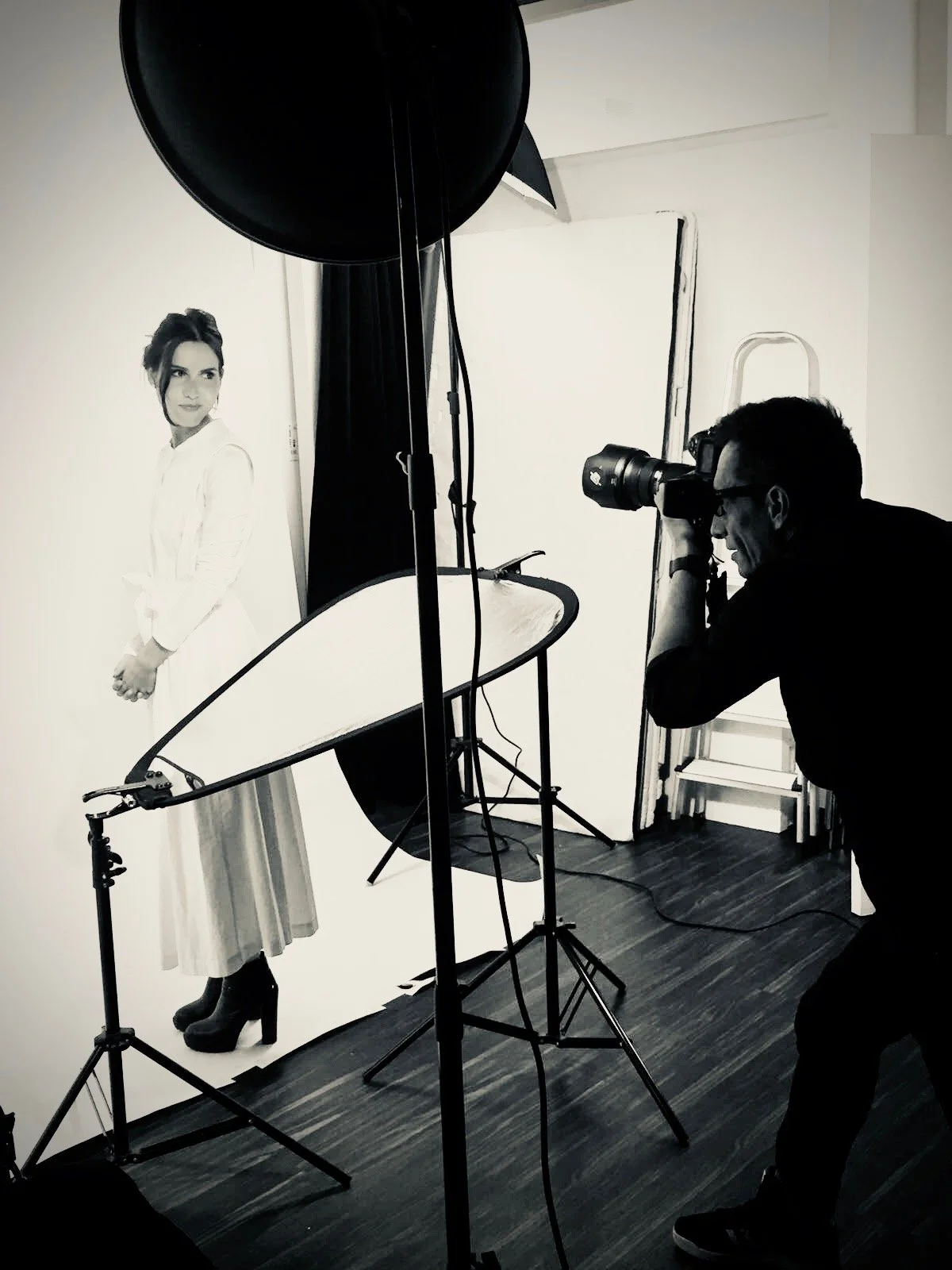 Une séance photo en studio avec un photographe prenant en photo une personne posant devant un fond blanc. Équipement d'éclairage et réflecteurs sont visibles.