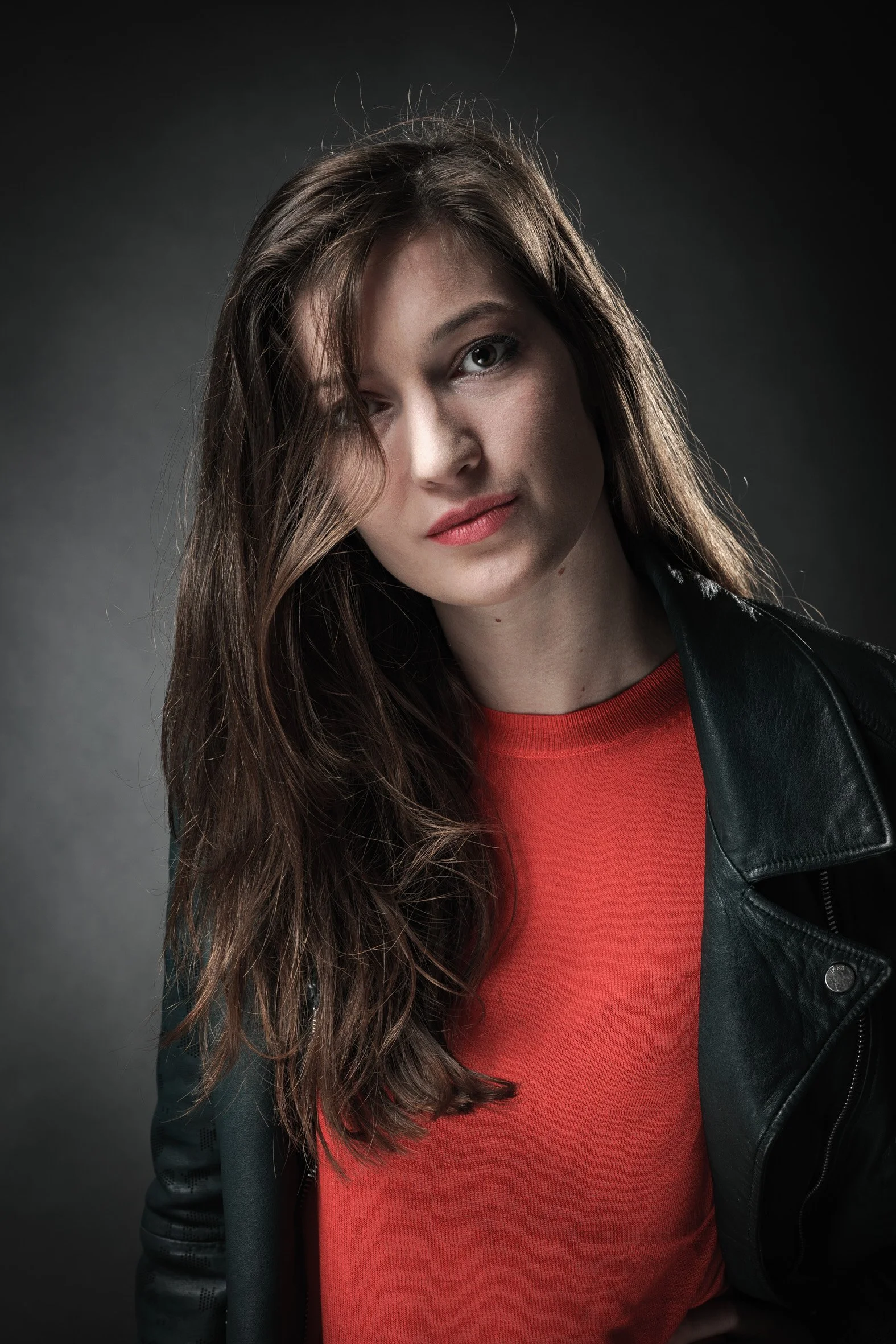 Une femme avec de longs cheveux bruns portant un pull rouge et une veste en cuir noir, posant devant un fond sombre.