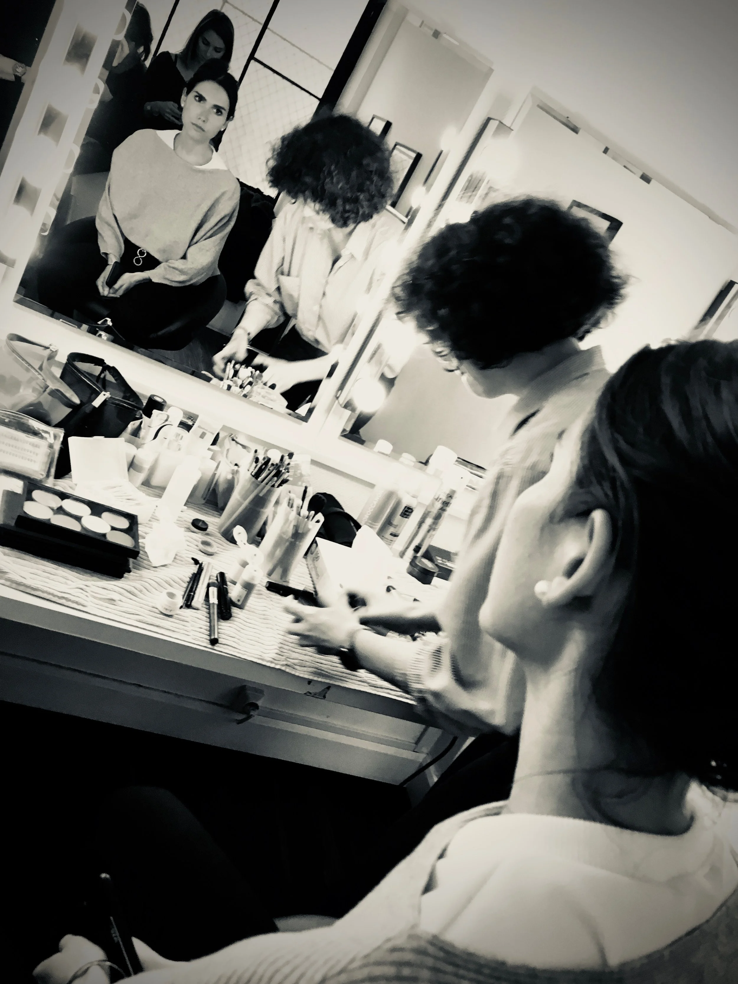 Deux personnes se préparent devant un miroir entouré de lumières, avec des produits de maquillage posés sur une table. Une personne est assise tandis qu'une autre semble en train de se maquiller. Reflets visibles dans le miroir.