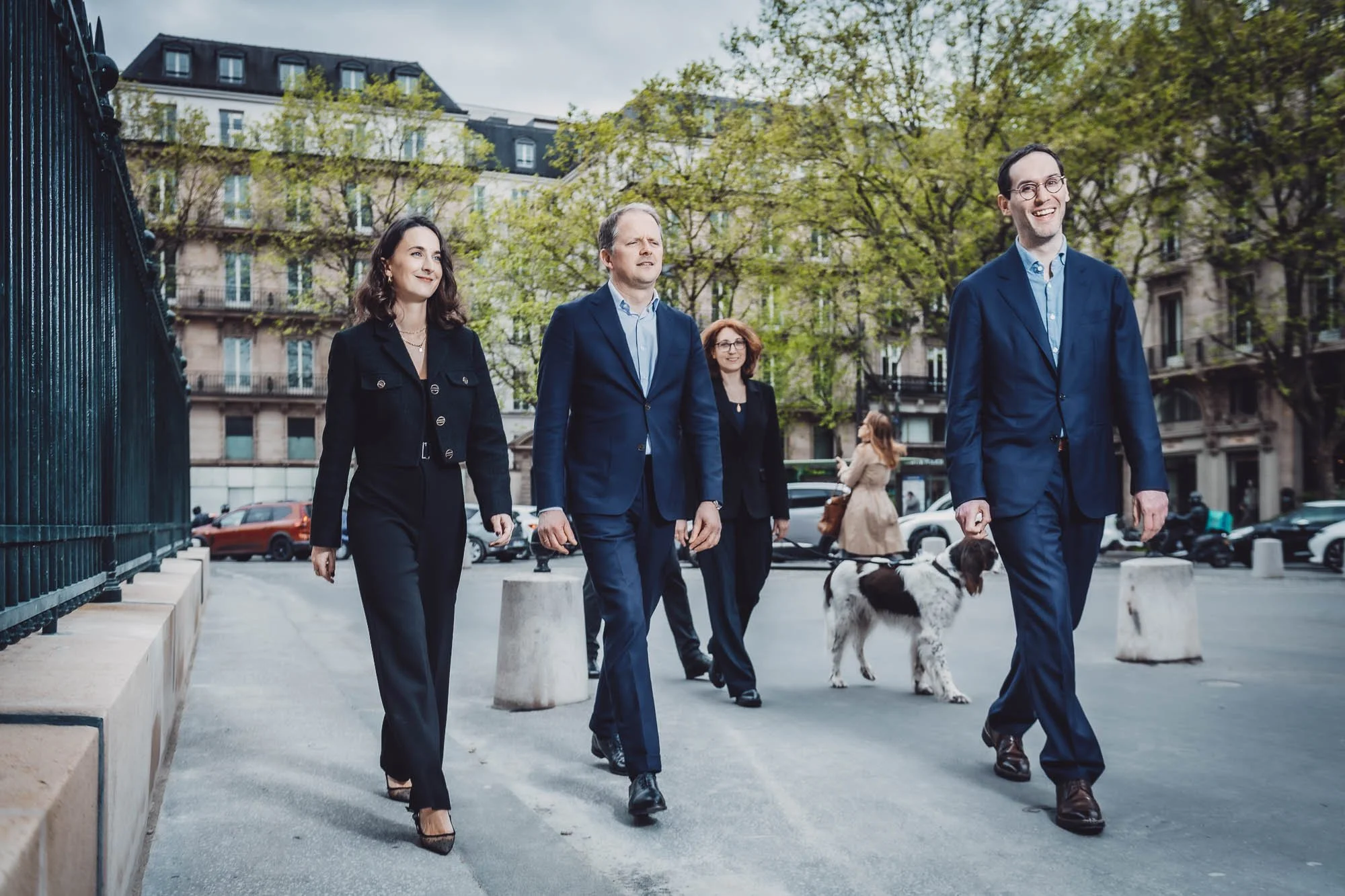 Quatre personnes en costume marchent dans une rue urbaine bordée d'arbres, avec des immeubles en arrière-plan.