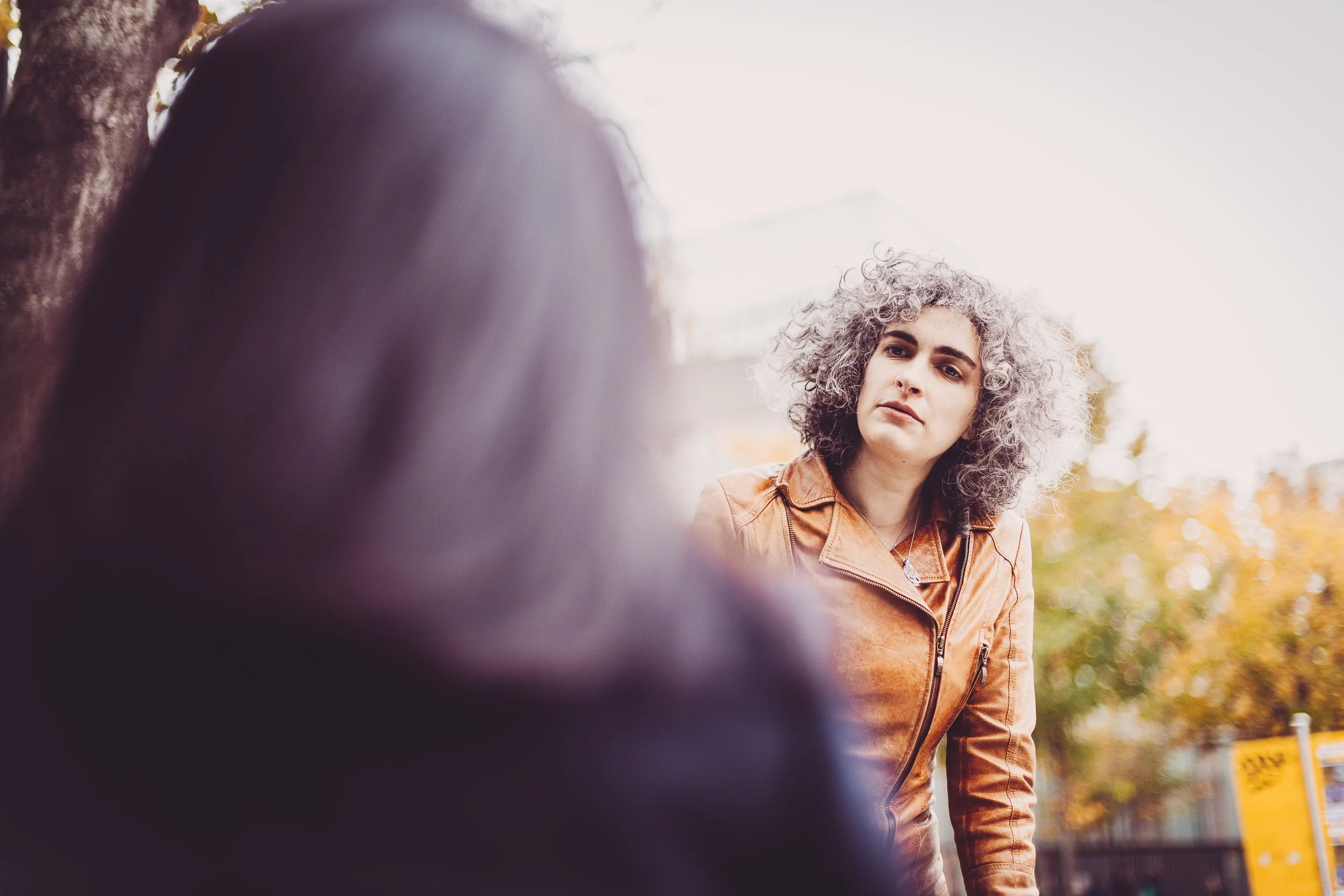 Personne aux cheveux gris portant une veste en cuir brun, regardant une autre personne floue à l'avant-plan, à l'extérieur avec des arbres en arrière-plan.