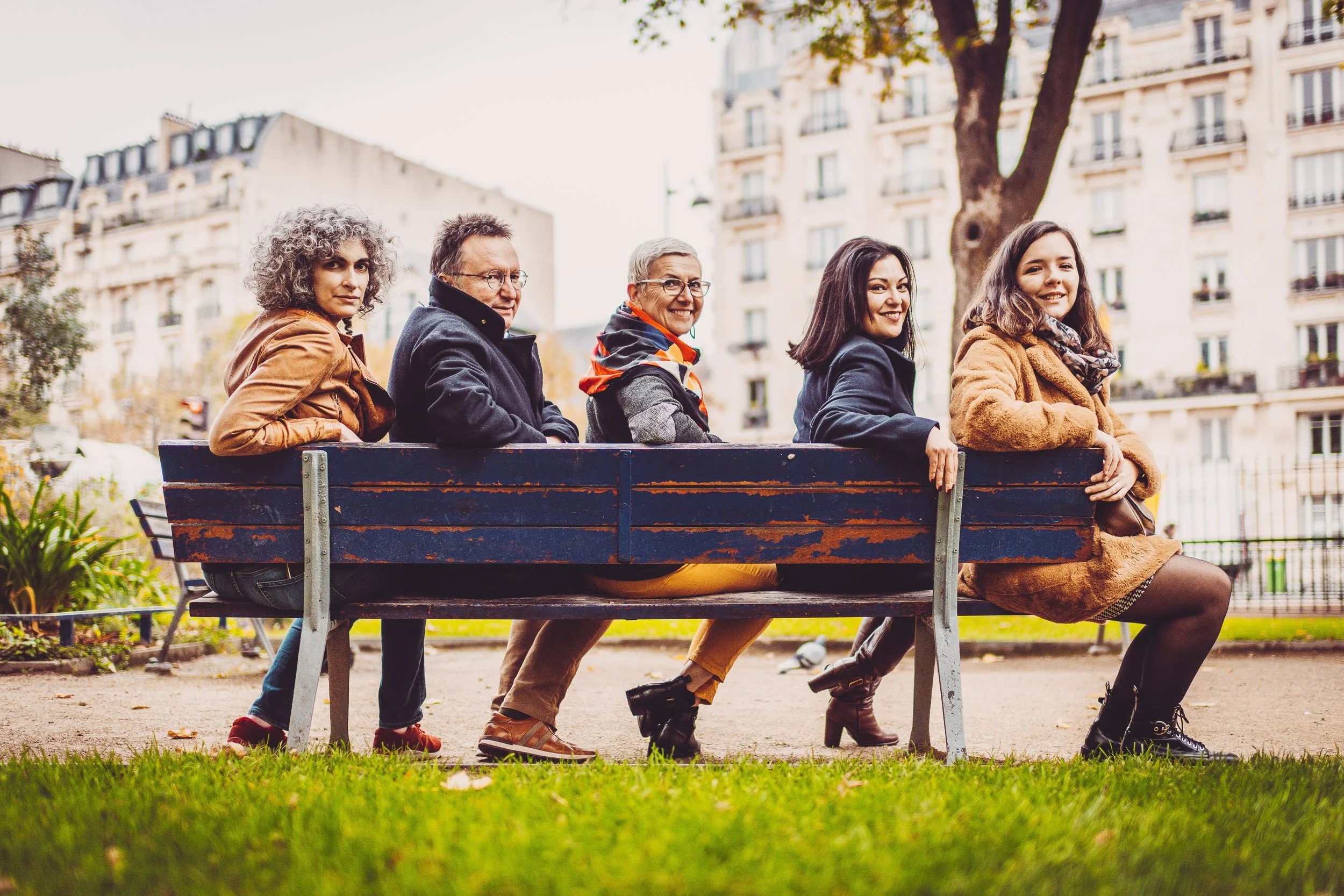 Cinq personnes assises sur un banc dans un parc urbain, souriantes. En arrière-plan, des bâtiments résidentiels.