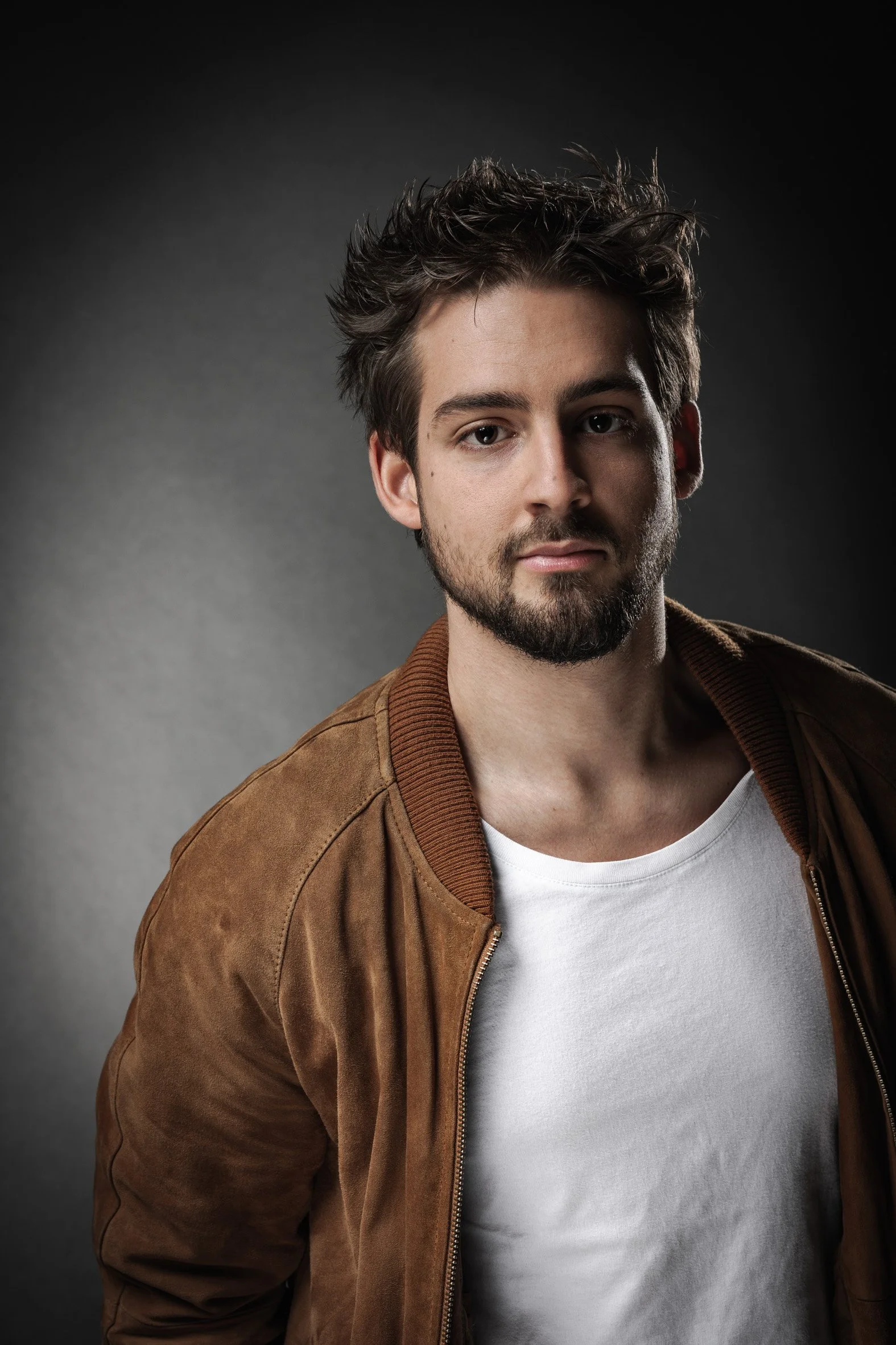 Un homme avec une barbe, portant un blouson marron et un t-shirt blanc, sur un fond gris.