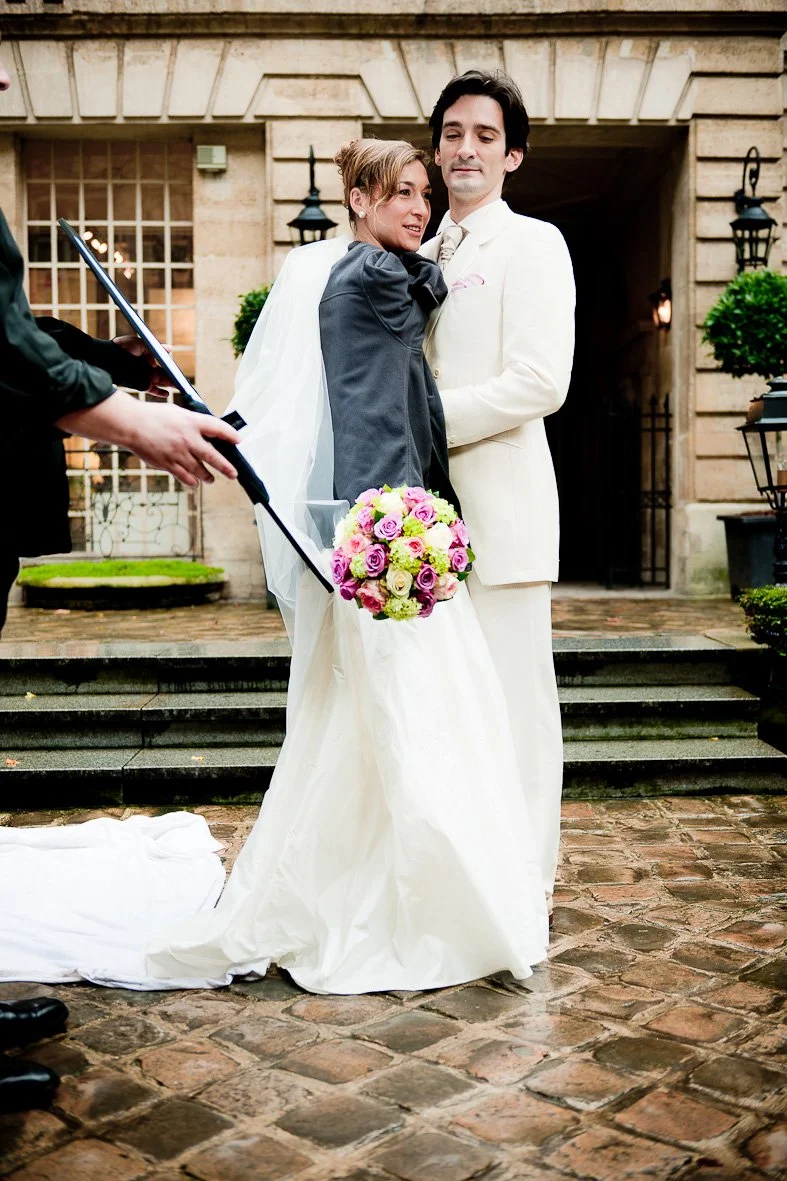 Un couple en tenue de mariage pose pour une photo en extérieur. La mariée porte une robe blanche et tient un bouquet de fleurs colorées. Le marié est en costume blanc. Une personne en costume rouge tient un parapluie au-dessus d'eux.