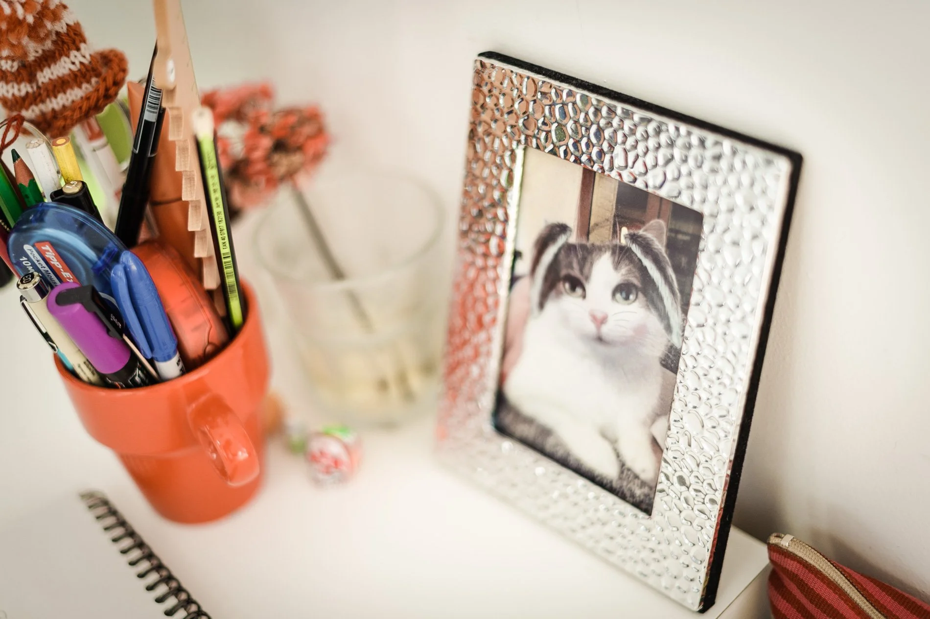 Un bureau avec une tasse rouge contenant des fournitures de bureau, un cadre photo avec une image de chat, un verre transparent et un carnet. Un bonbon est posé sur la table.