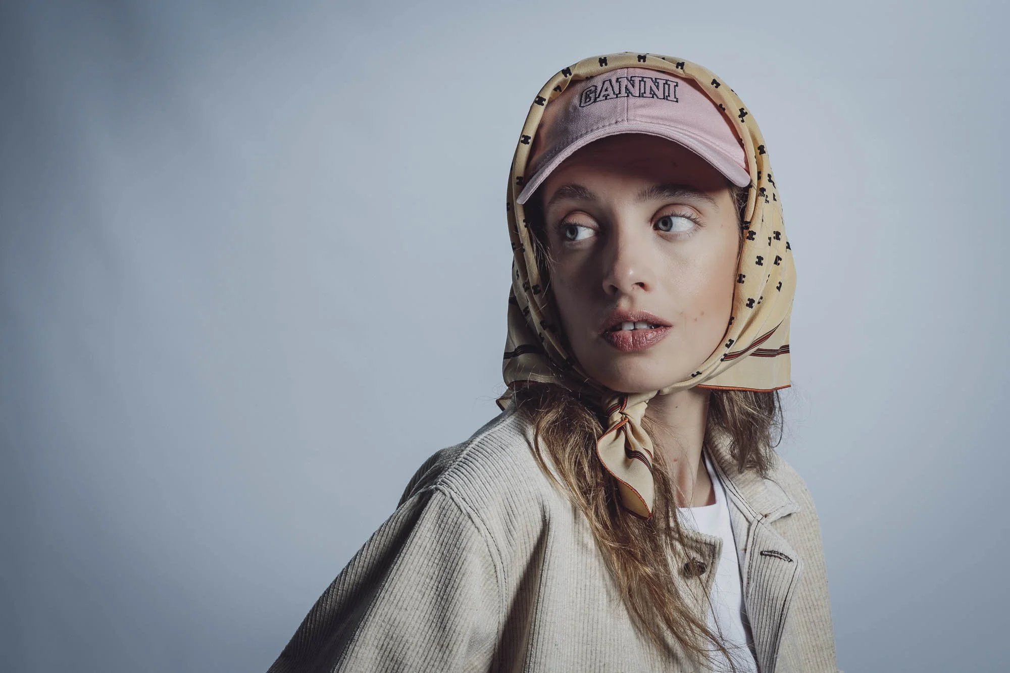 Femme portant un foulard jaune à motifs et une casquette rose, avec une veste beige et un fond flou gris.