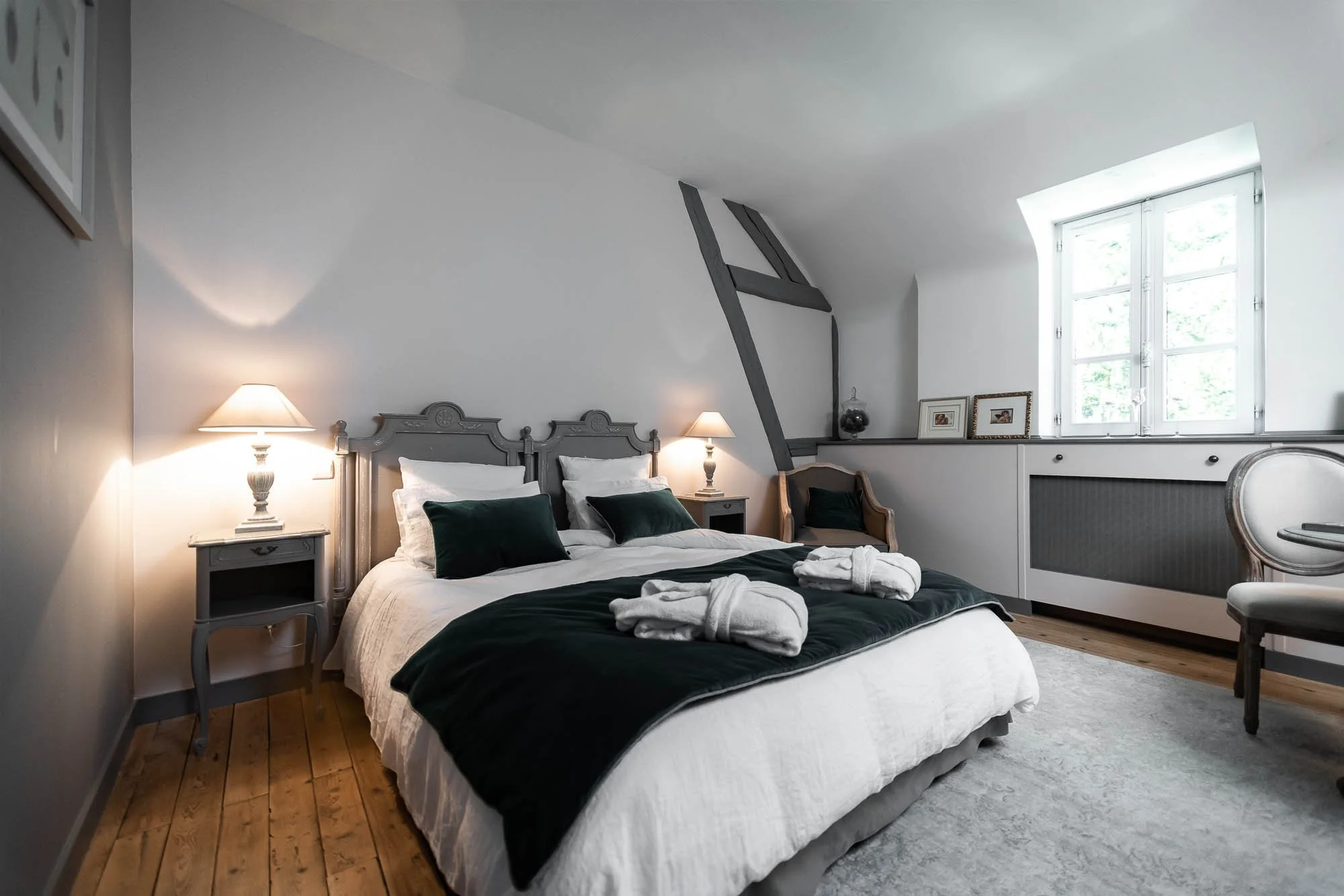 Chambre élégante avec un lit double, draps blancs, coussins verts, deux lampes de chevet, une chaise et une fenêtre.