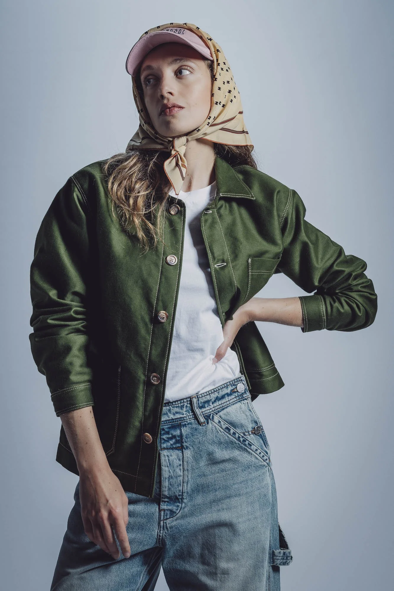 Femme portant une veste verte, un foulard à motifs et une casquette rose, posant avec une main sur la hanche et un jean décontracté.