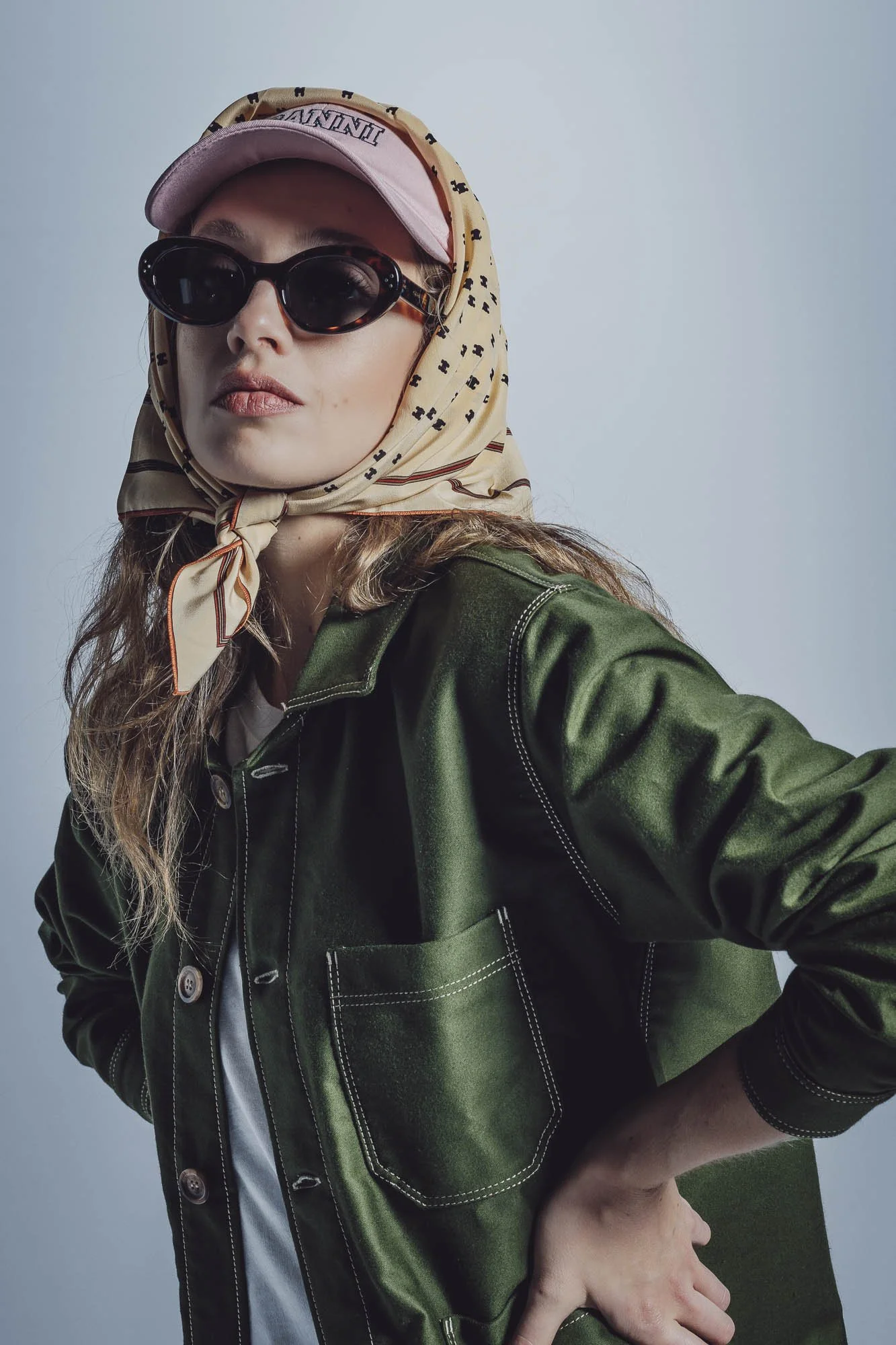 Femme portant une casquette rose, lunettes de soleil et un foulard, vêtue d'une veste verte.