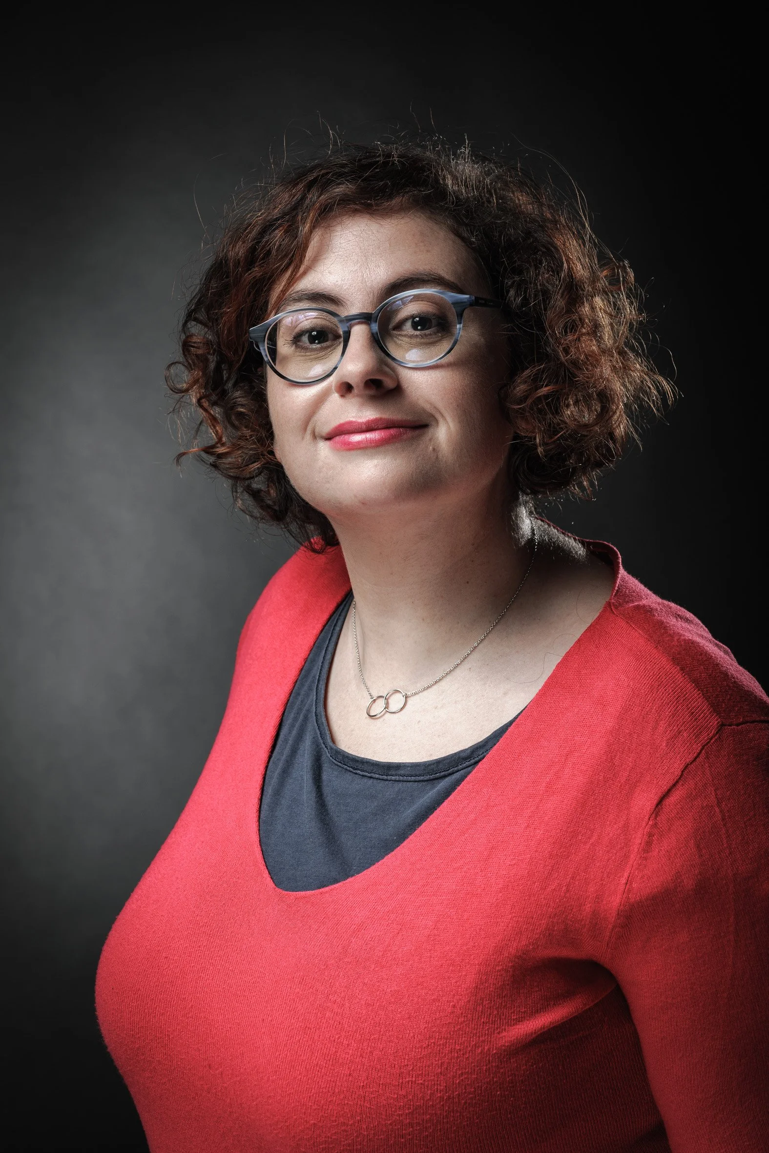 Une femme avec des cheveux courts et bouclés, portant des lunettes et un pull rouge, sourit légèrement devant un fond sombre. Elle porte un collier avec un pendentif en forme de cercles entrelacés.
