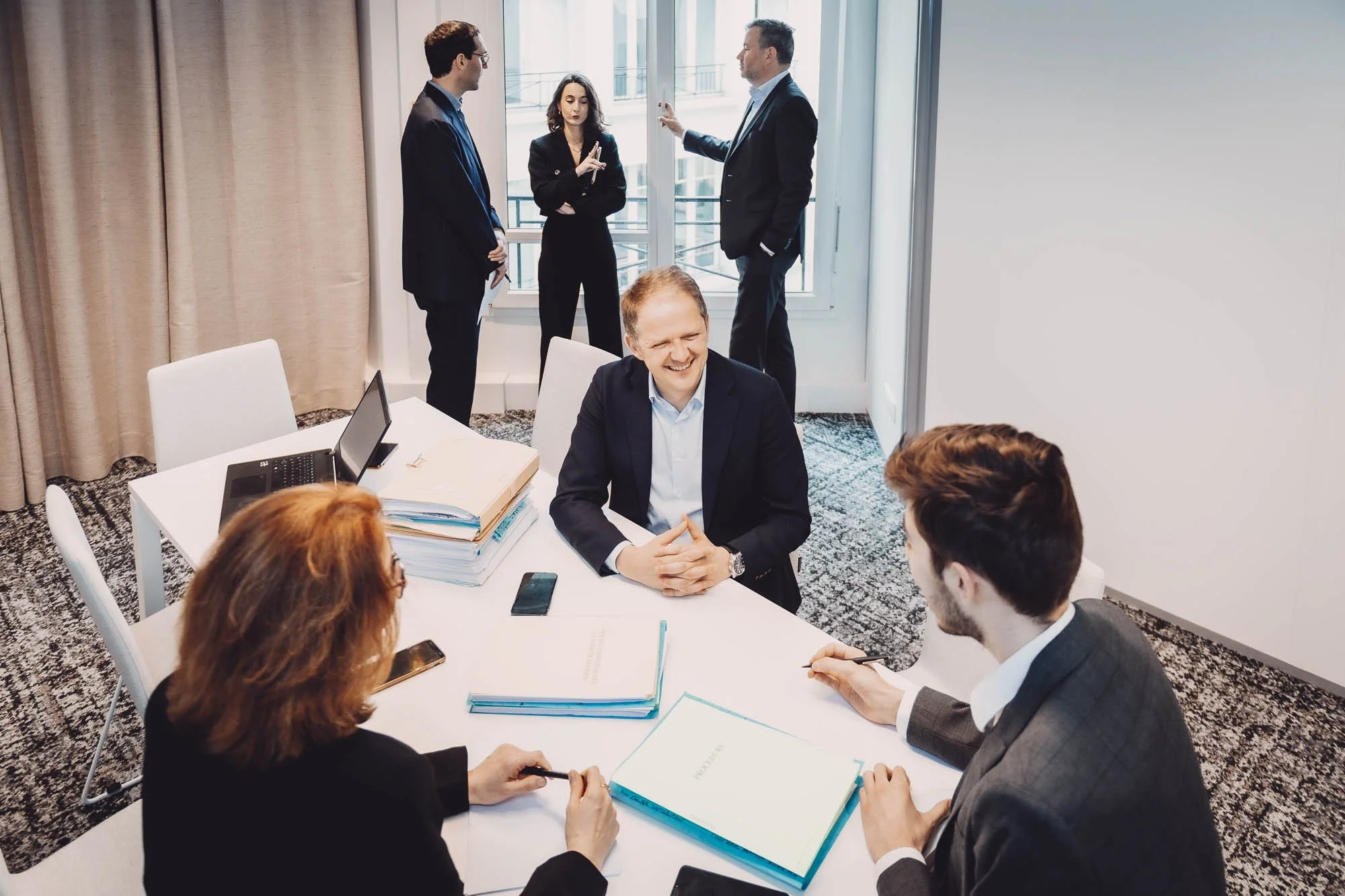 Réunion de bureau avec plusieurs personnes en costumes discutant autour d'une table avec des dossiers, un ordinateur portable, et une ambiance professionnelle.