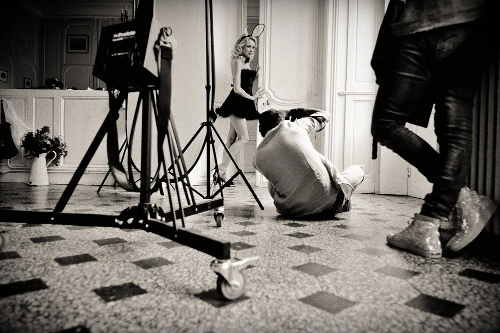 Séance photo en cours avec un photographe prenant une photo d'une femme portant des oreilles de lapin et une robe noire. Deux autres personnes sont présentes avec de l'équipement photographique. Image en noir et blanc.