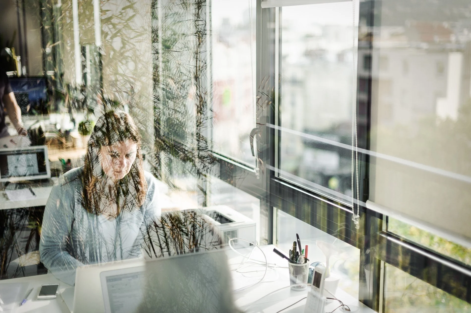 Une femme travaille sur un ordinateur portable dans un bureau moderne avec une grande fenêtre donnant sur un paysage urbain.