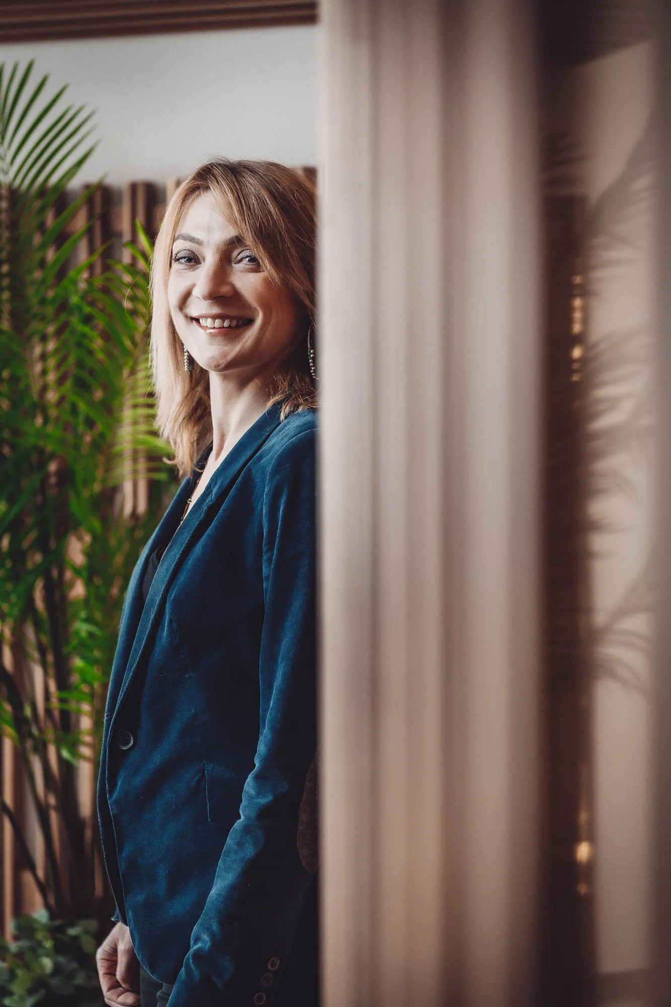 Femme souriante en blazer bleu, debout près d'une plante verte.