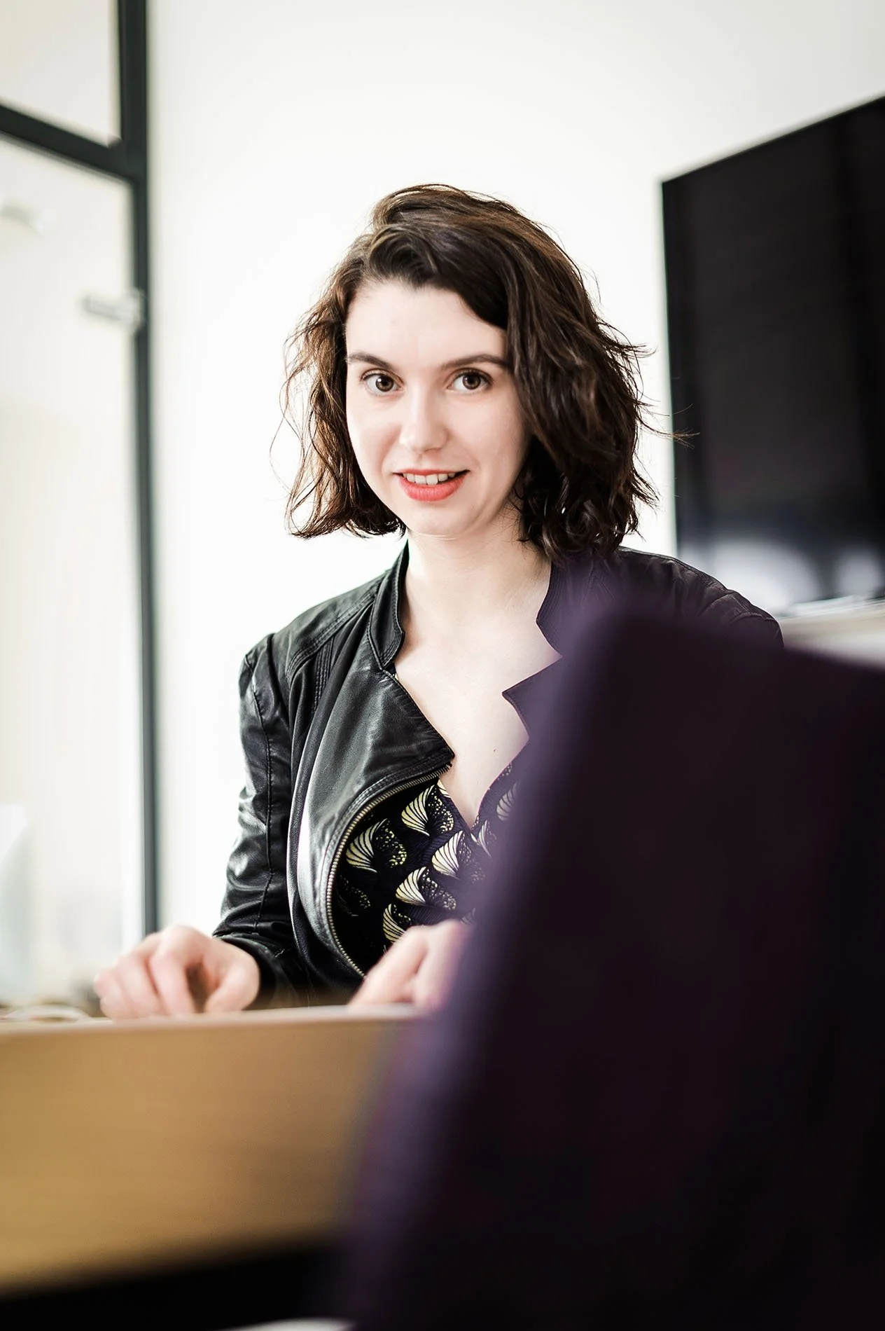Une femme avec des cheveux bruns courts, portant une veste en cuir noir, est assise devant un ordinateur dans un bureau lumineux.