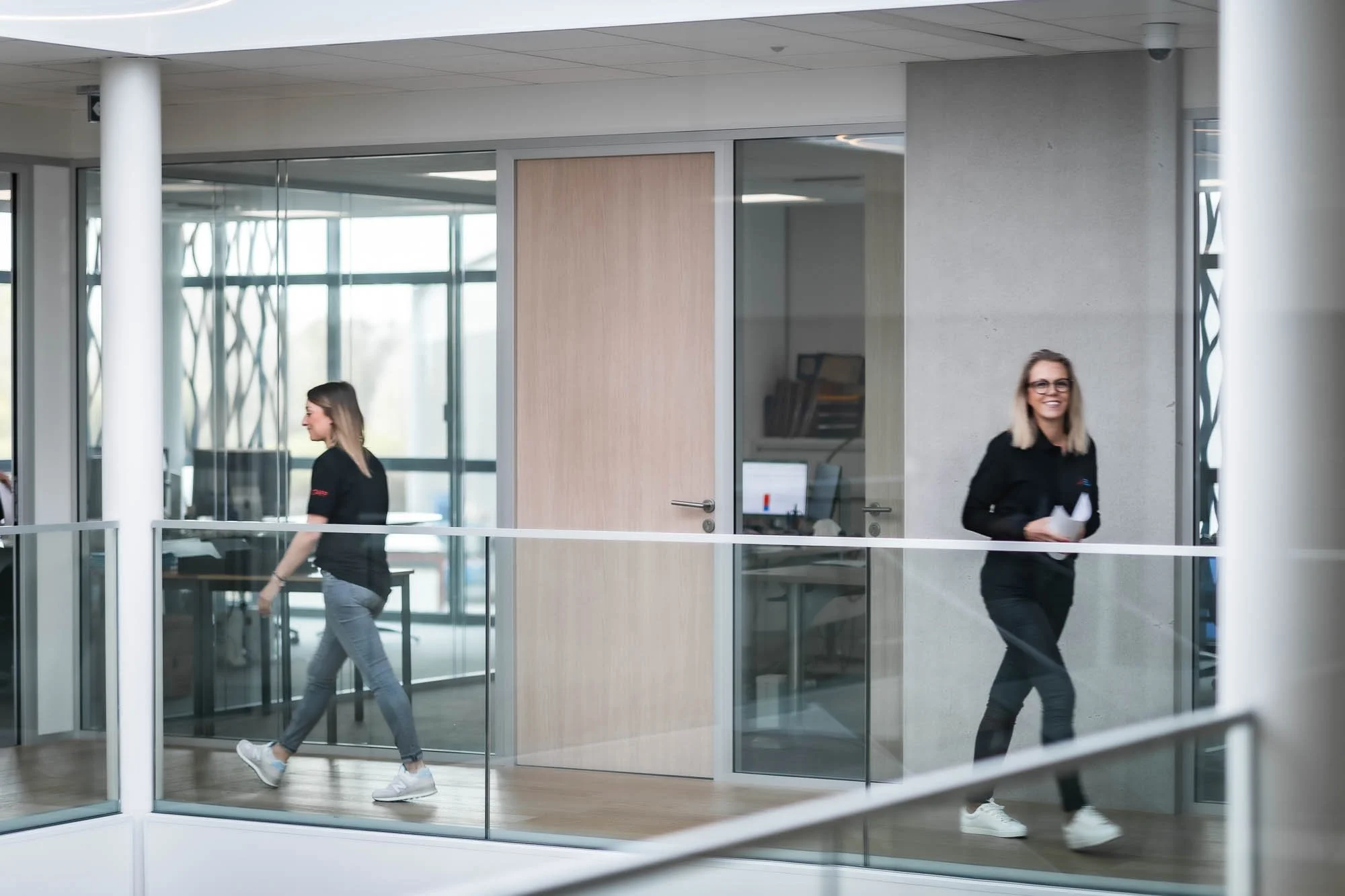 Deux femmes marchent dans un couloir de bureau moderne avec des murs de verre et une porte en bois clair.