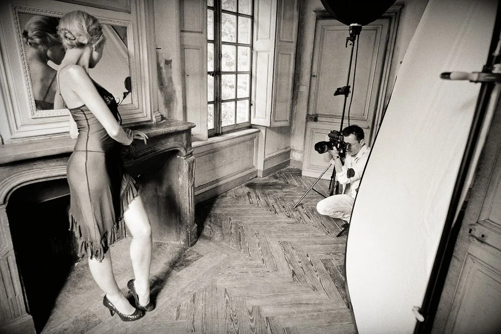 Séance photo en intérieur avec une femme en robe posant devant un photographe dans une pièce avec parquet et grandes fenêtres.