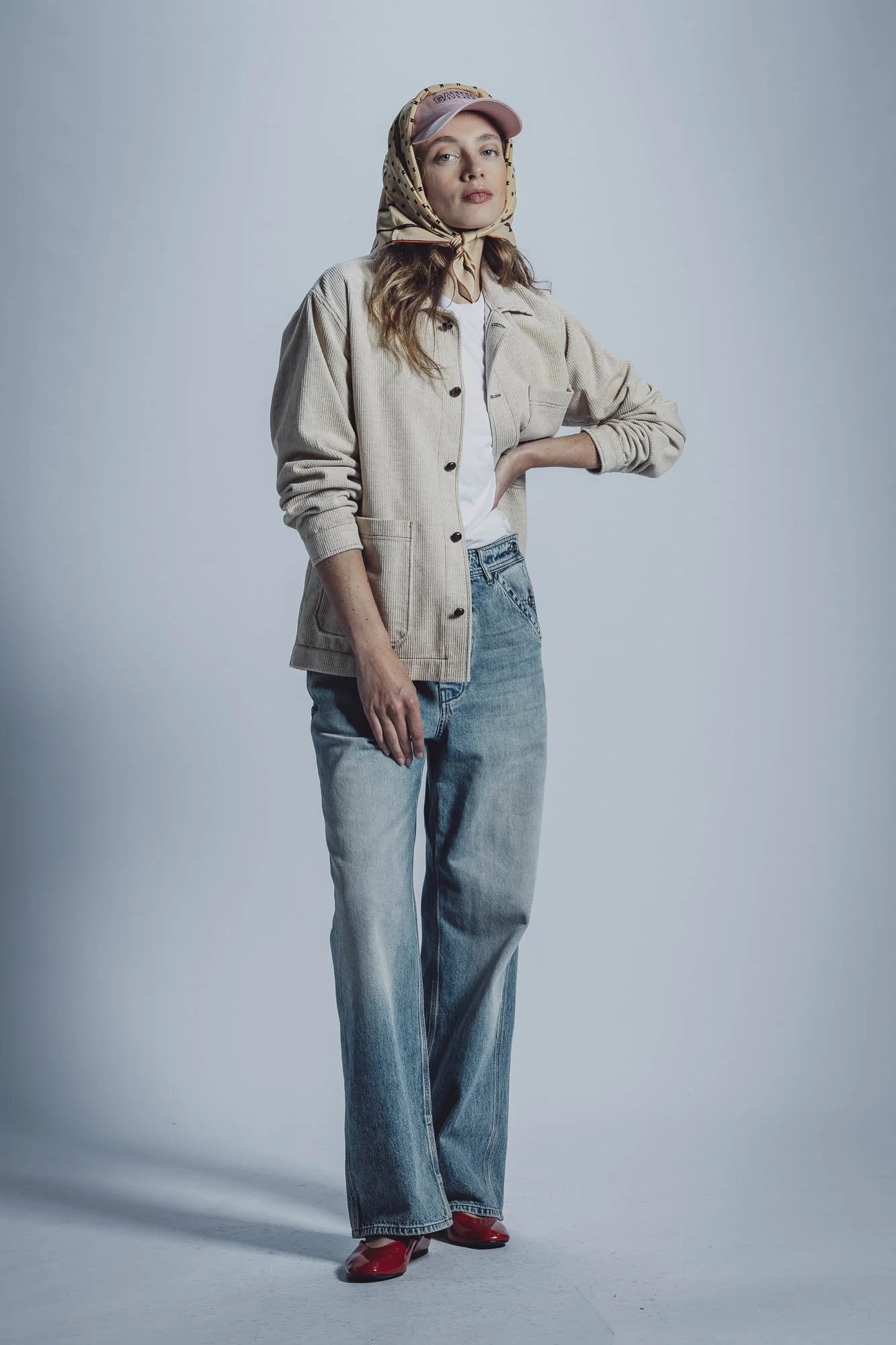 Femme portant un jean large, une veste beige, un t-shirt blanc sous la veste, un foulard sur la tête, une casquette rose et des chaussures rouges. Elle pose debout, une main sur la hanche.