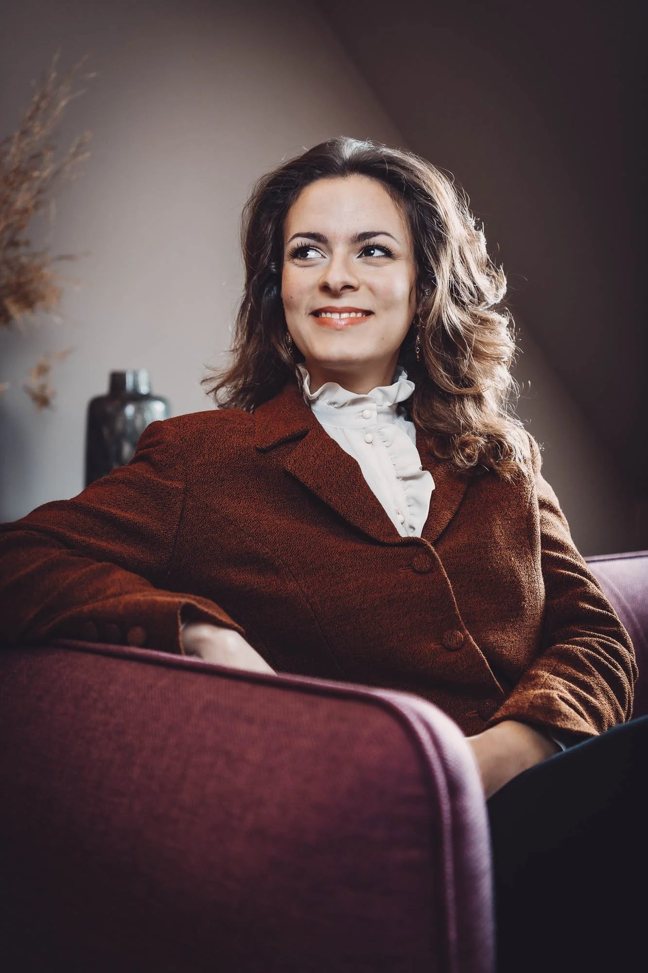 Une femme souriante assise sur un canapé, portant une veste marron et une chemise blanche avec des éléments de décoration en arrière-plan.