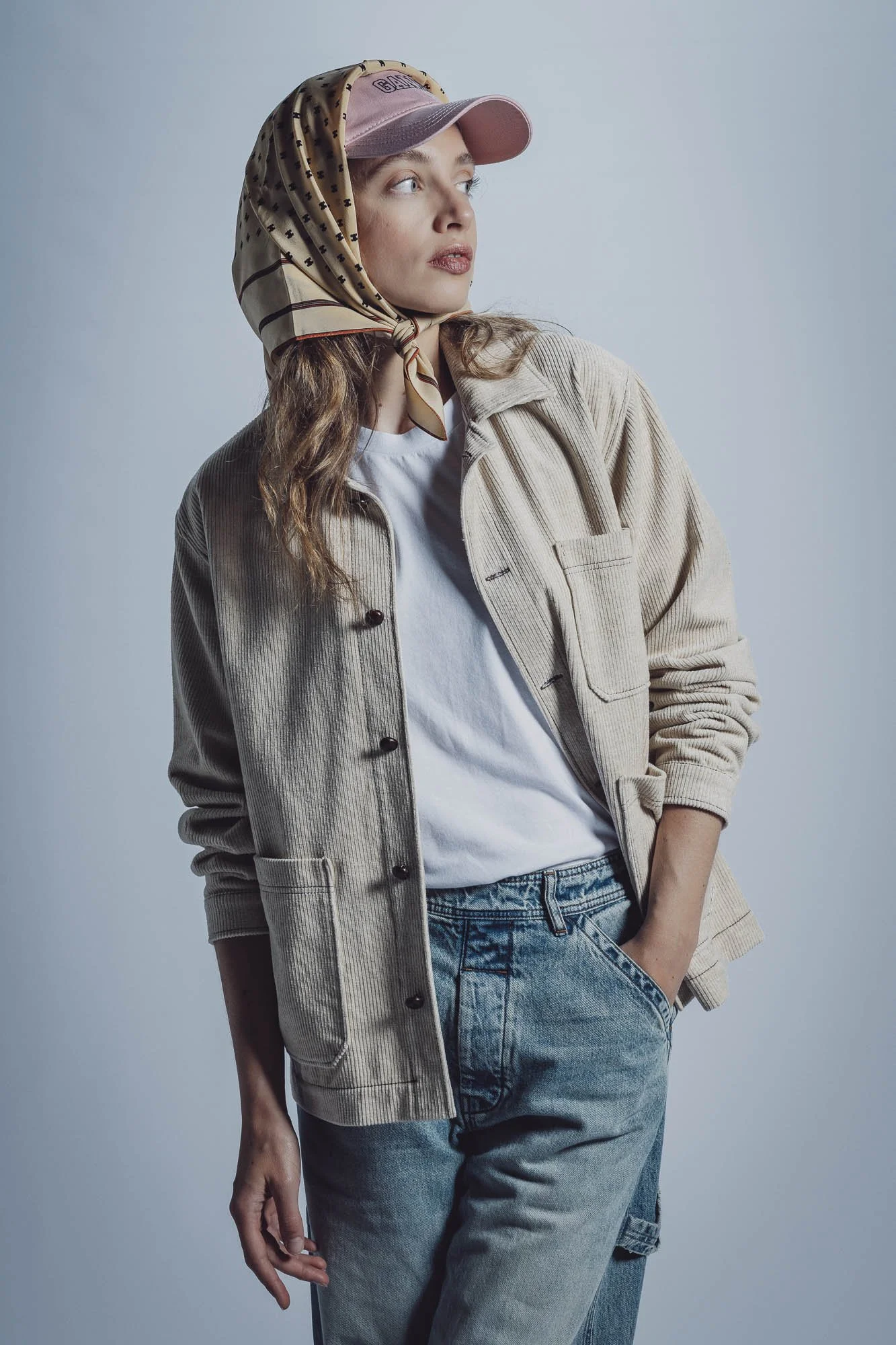 Femme portant une casquette rose et un foulard, vêtue d'une veste beige, un t-shirt blanc et un jean, regardant sur le côté.