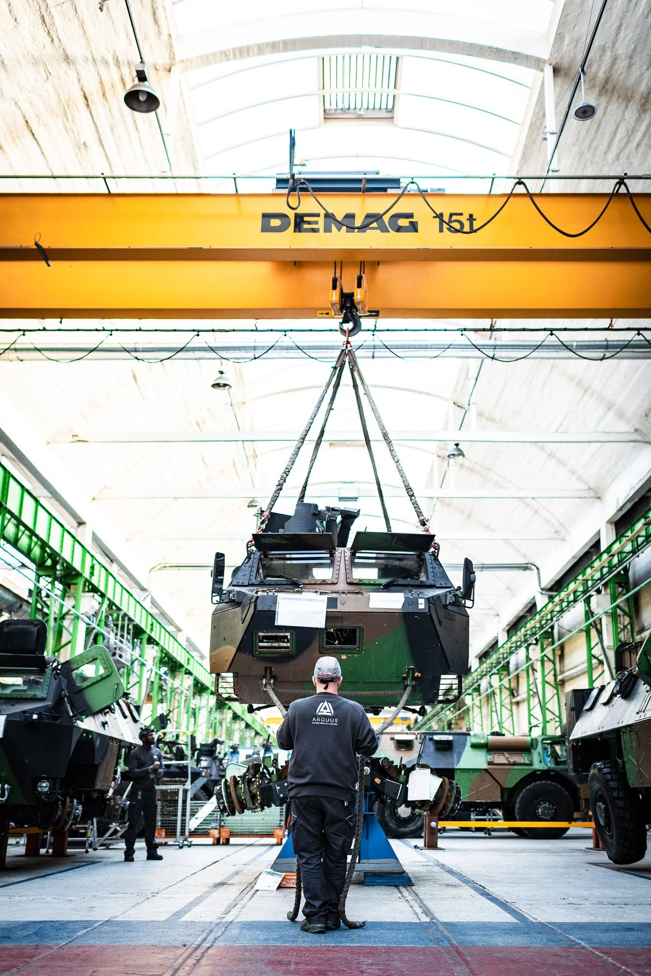 Un véhicule militaire en cours de fabrication dans une usine, soulevé par une grue Demag, avec des ouvriers travaillant autour.