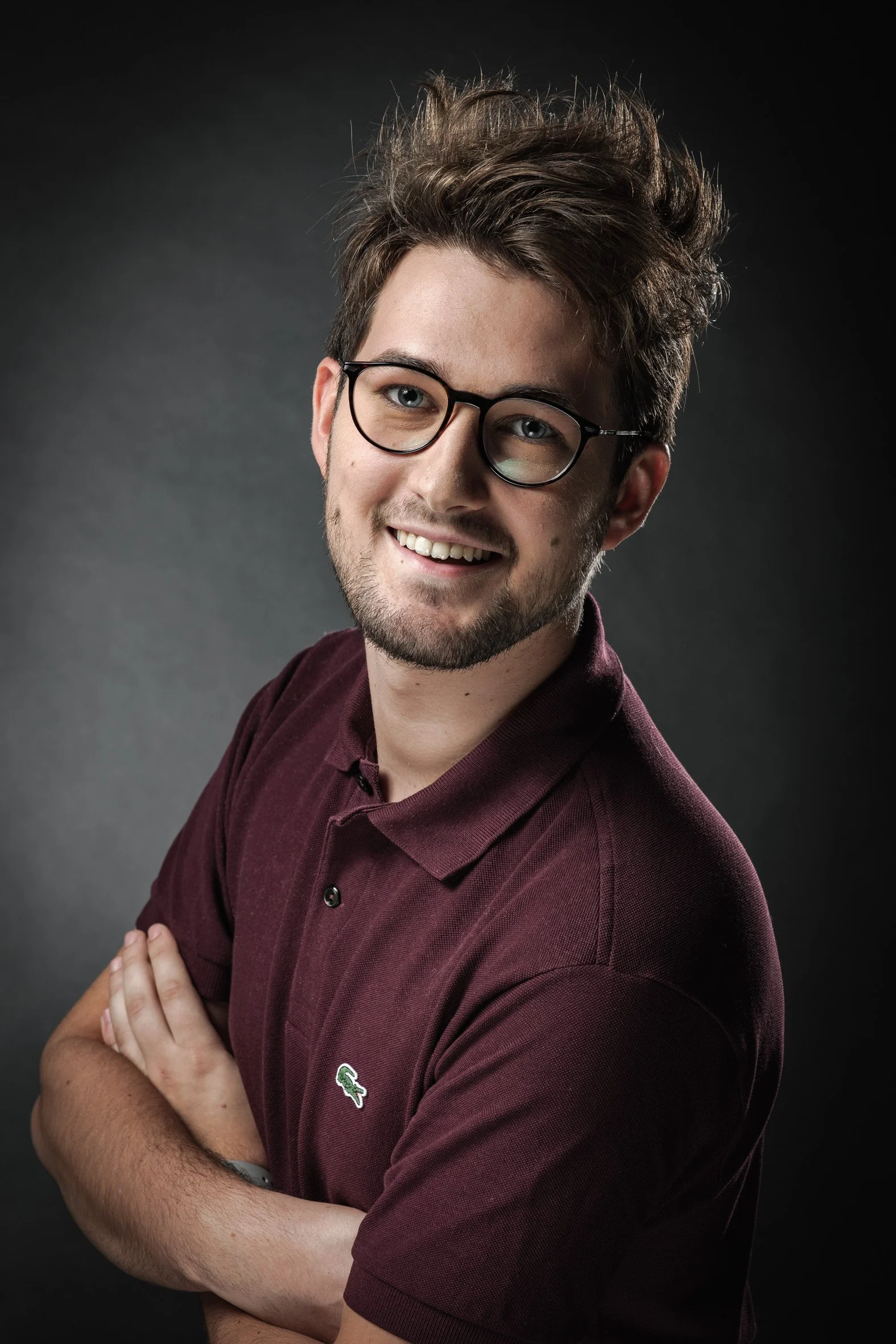 Un homme souriant avec des lunettes et une barbe, portant un polo bordeaux, sur un fond sombre.