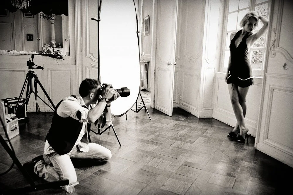 Un photographe prend une photo d'un modèle féminin posé près d'une fenêtre, dans un studio éclairé par des équipements de photographie.
