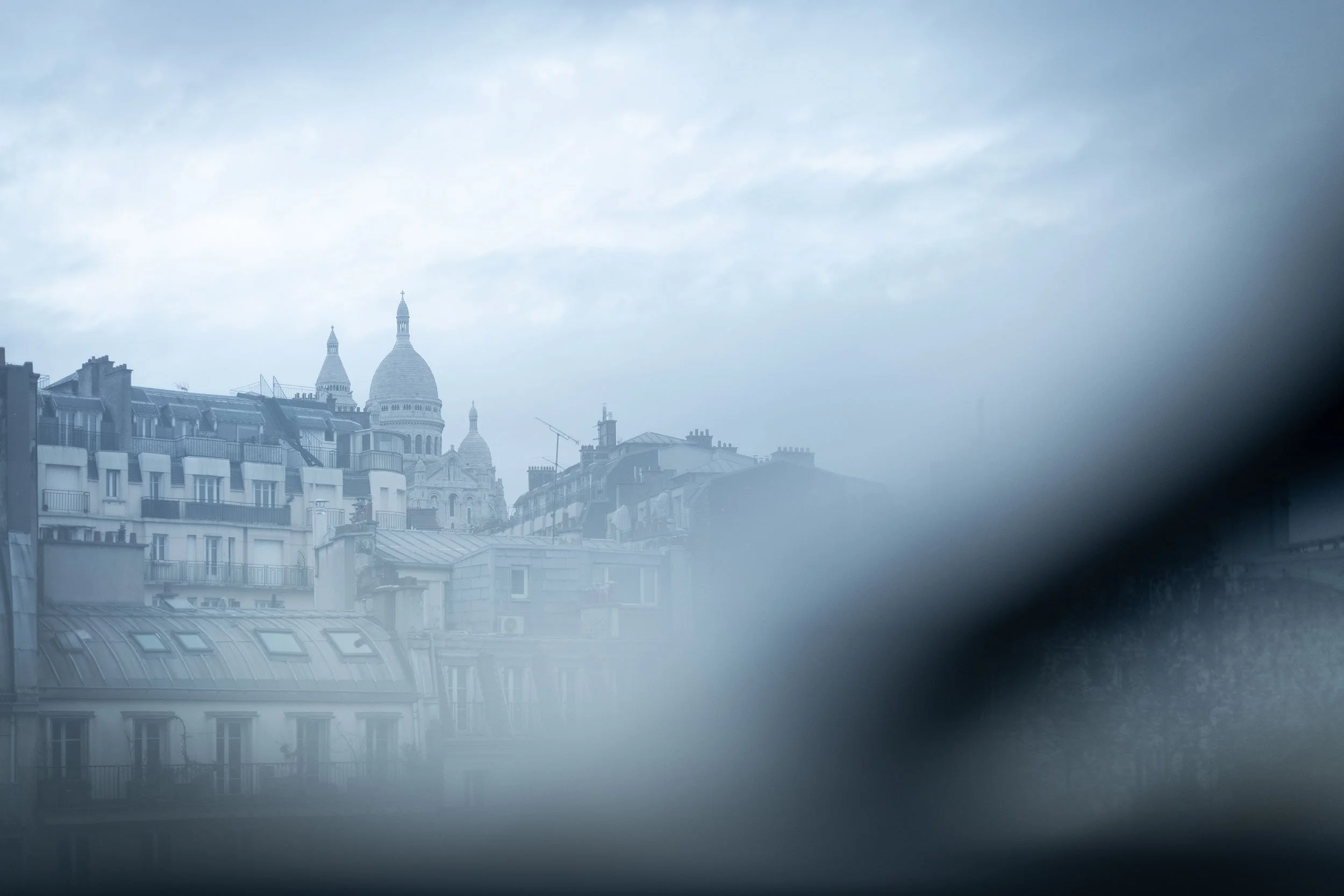 Vue floue de la basilique du Sacré-Cœur à Paris derrière des immeubles sous un ciel nuageux.