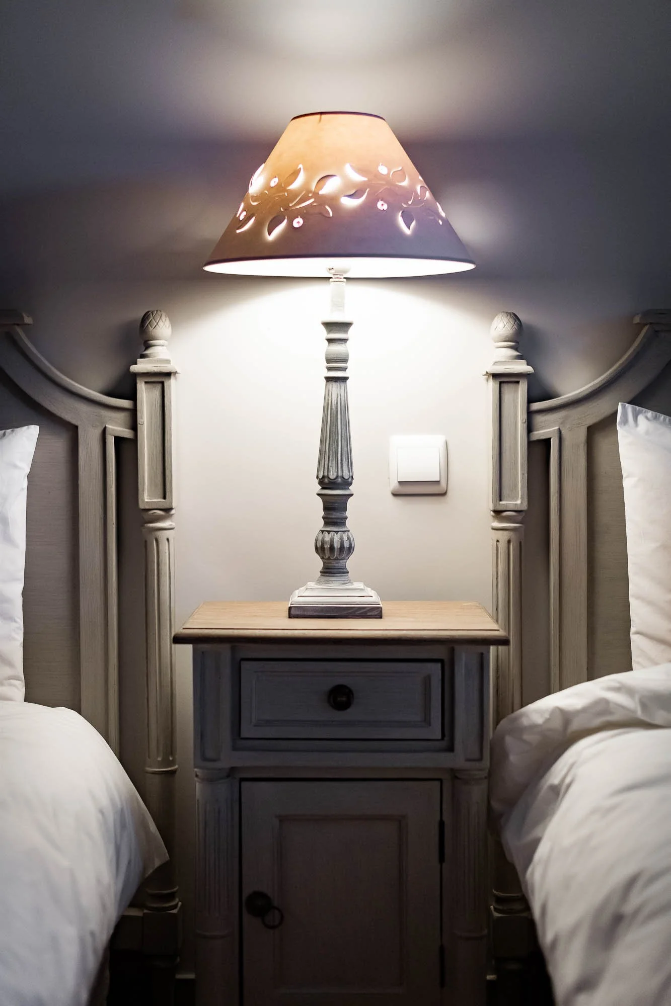 Lampe de chevet allumée sur une table de nuit en bois entre deux lits, avec des têtes de lit en bois sculpté.