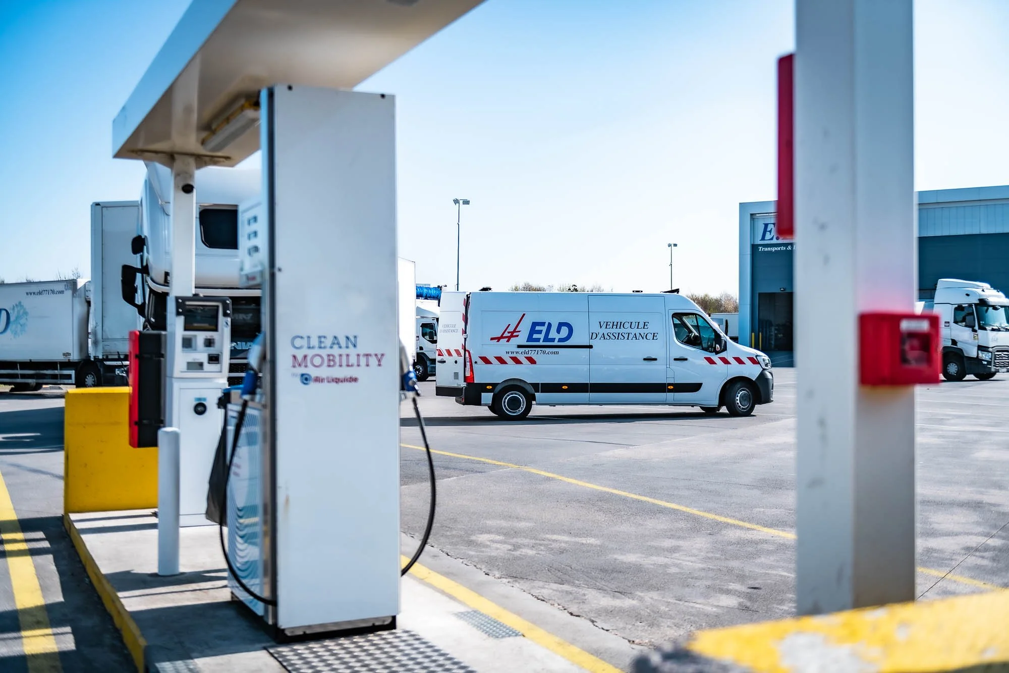 Une station de ravitaillement de carburant avec une pompe de "Clean Mobility" au premier plan. À l'arrière-plan, une camionnette d'assistance "ELD" et des camions stationnés.