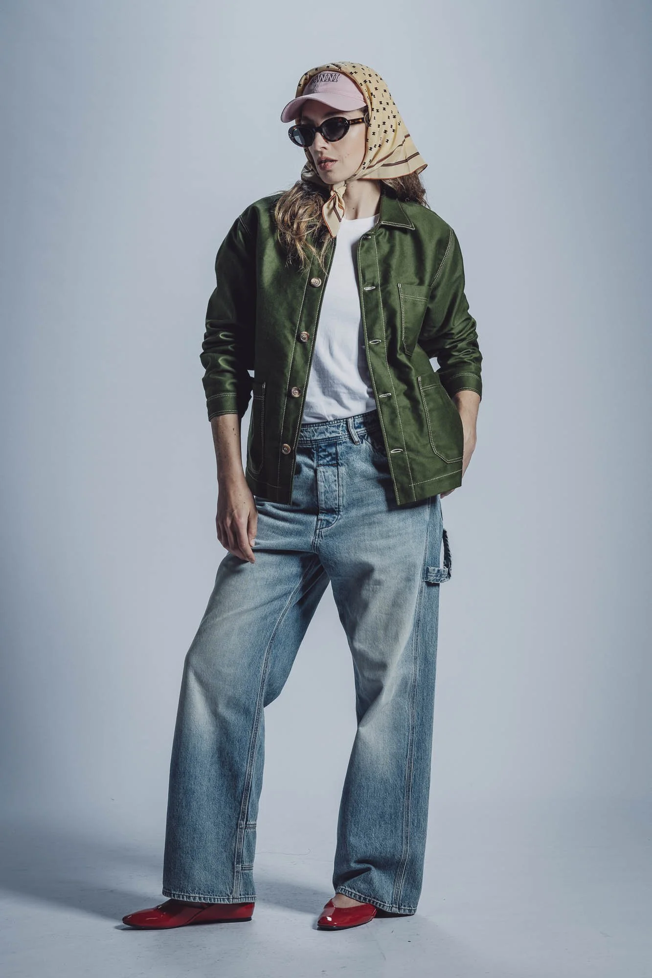 Femme portant une casquette rose, lunettes de soleil, foulard jaune, veste vert foncé, jean ample et chaussures rouges.