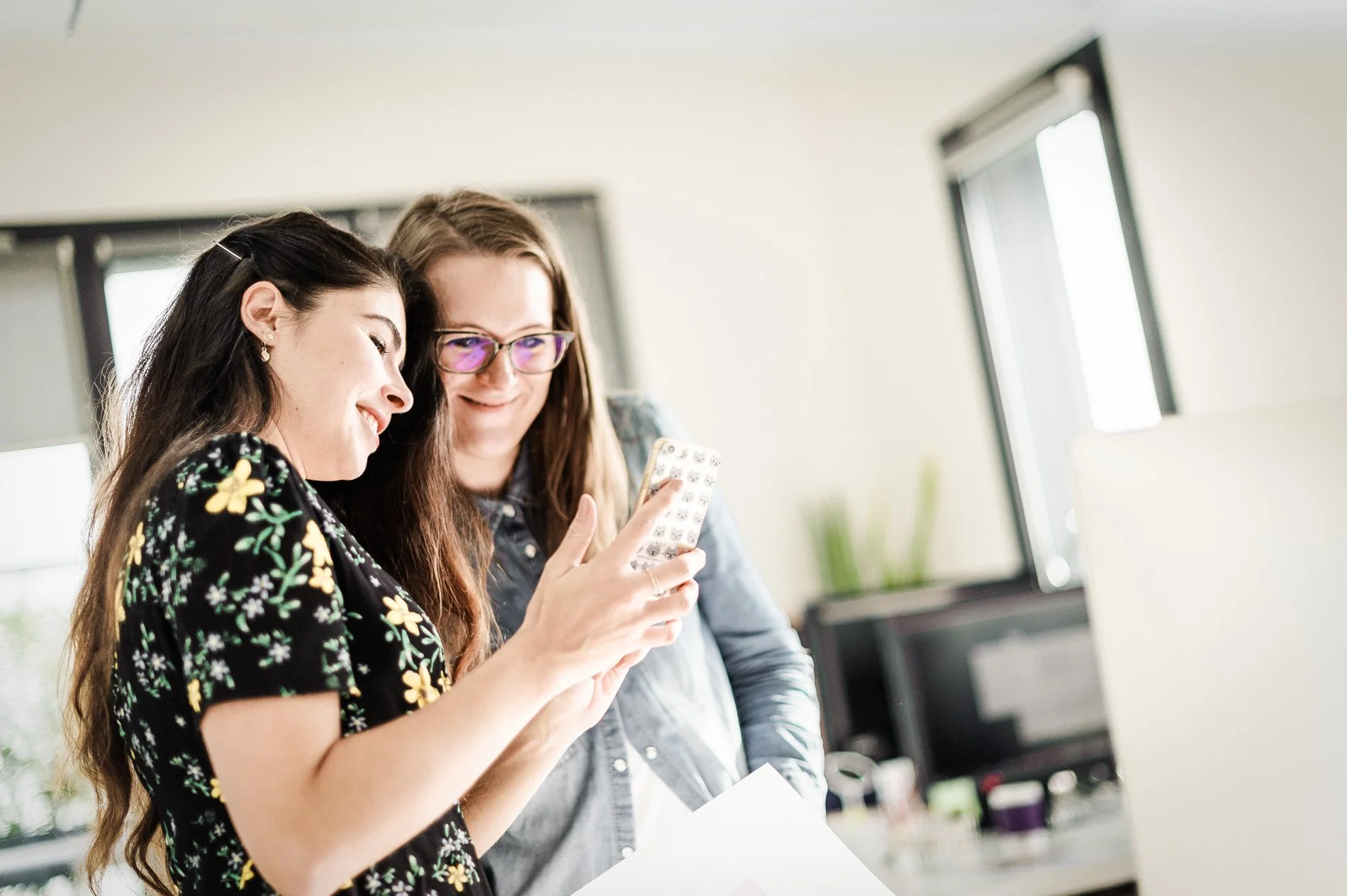 Deux femmes souriantes regardent un téléphone ensemble dans un bureau lumineux.