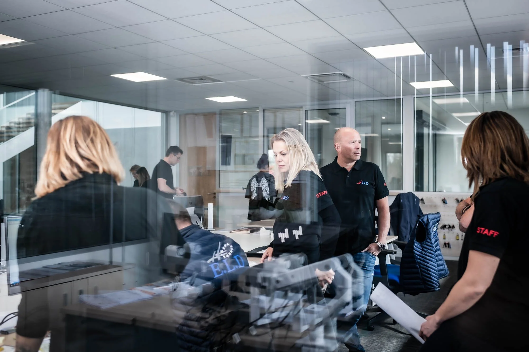 Personnes discutant dans un bureau moderne, partiellement vu à travers une vitre, portant des chemises avec "Staff" écrit dessus.