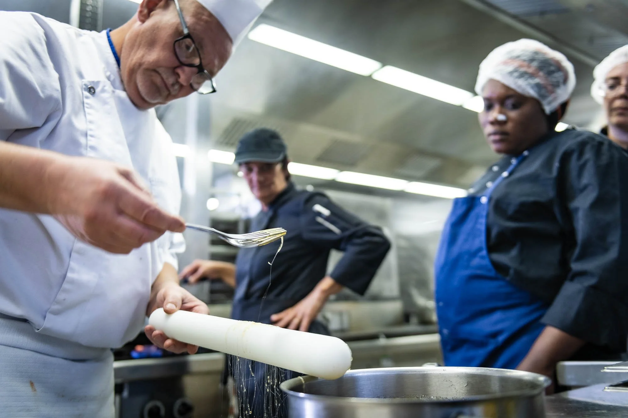 Un chef cuisinier versant un ingrédient avec une fourchette dans une cuisine, observé par deux autres personnes portant des tenues de cuisine.