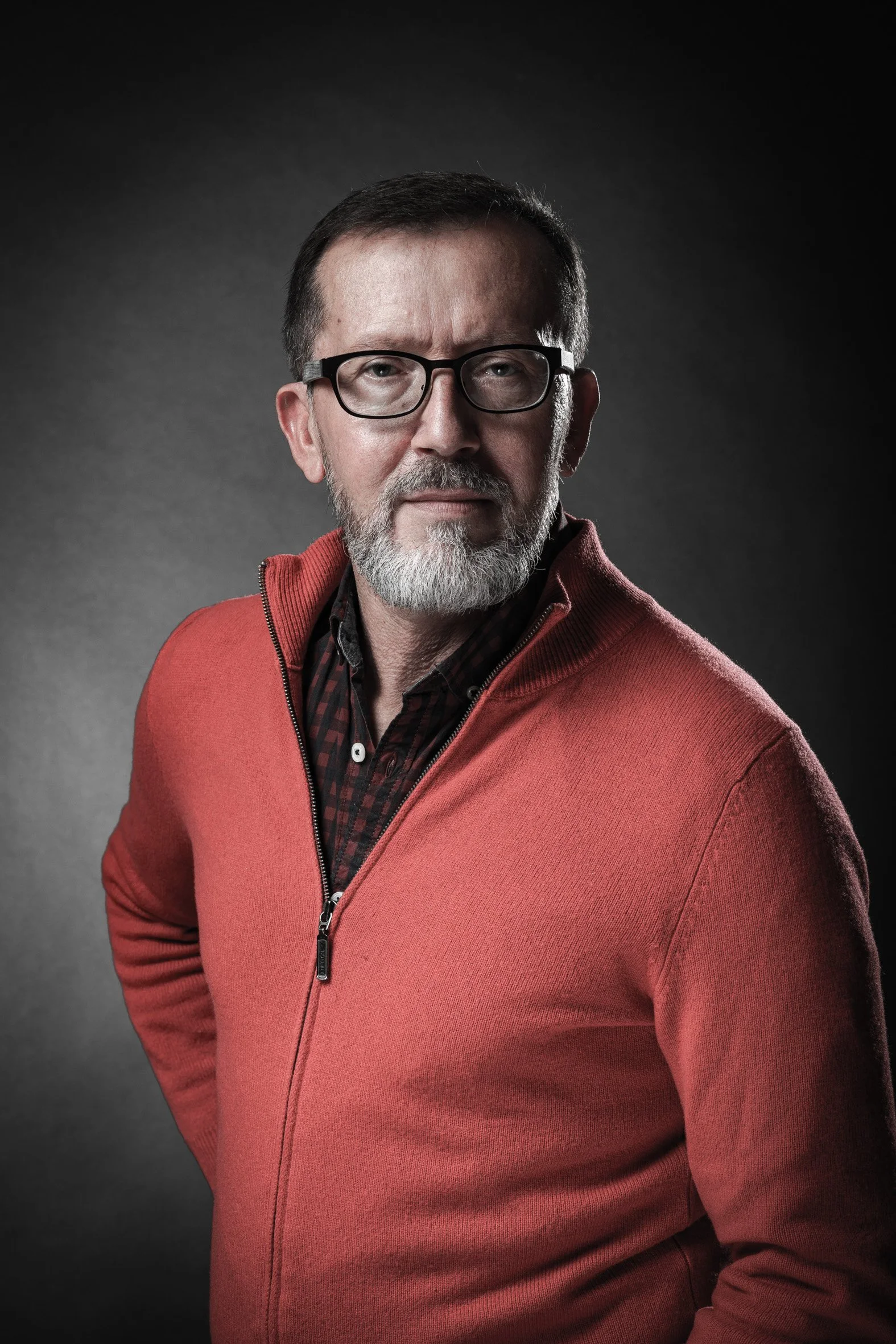 Portrait d'un homme portant des lunettes, une barbe et un pull rouge, avec une expression sérieuse.