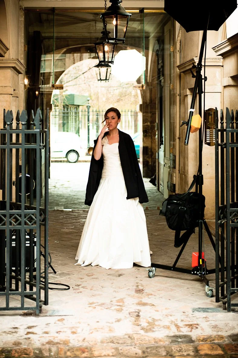 Une femme en robe de mariée blanche, avec un manteau noir, fume une cigarette dans une rue ou un passage couvert avec un éclairage et équipement photographique autour.