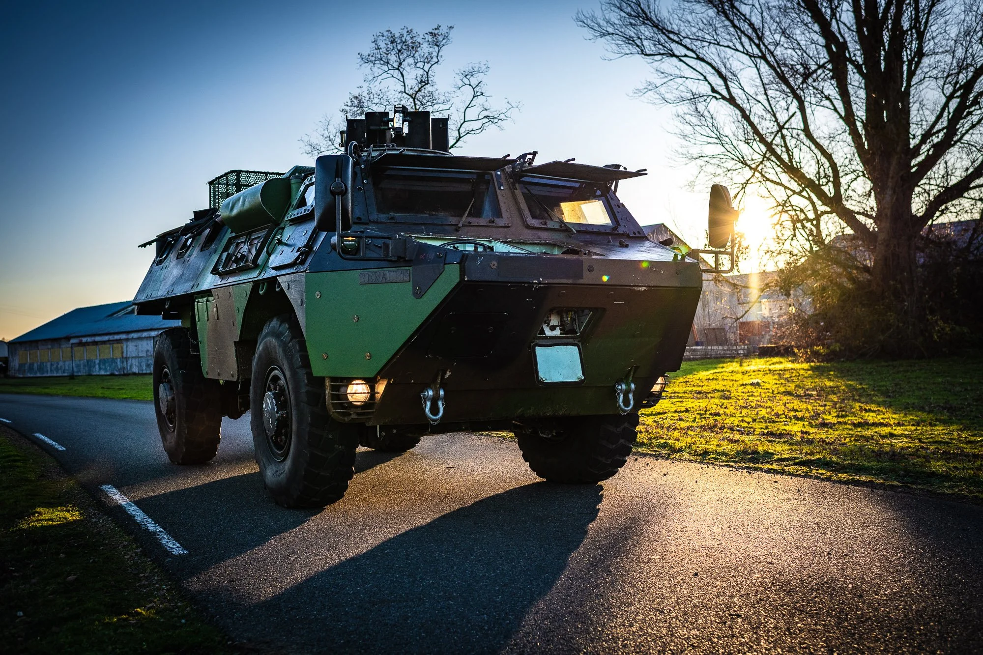 Véhicule blindé militaire sur une route avec des arbres en arrière-plan et le soleil couchant.