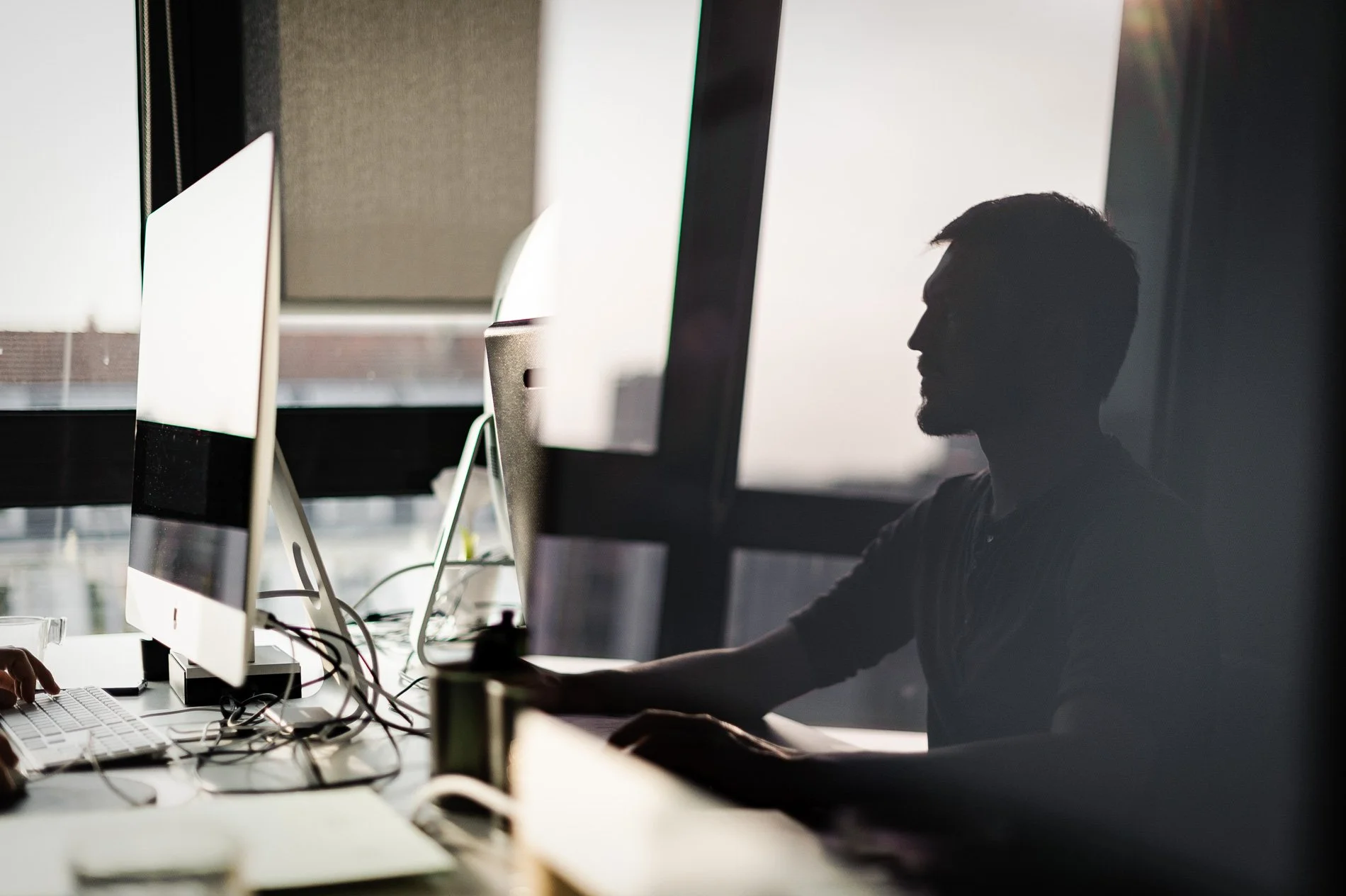 Silhouette d'un homme travaillant sur un ordinateur dans un bureau bien éclairé avec de grandes fenêtres.