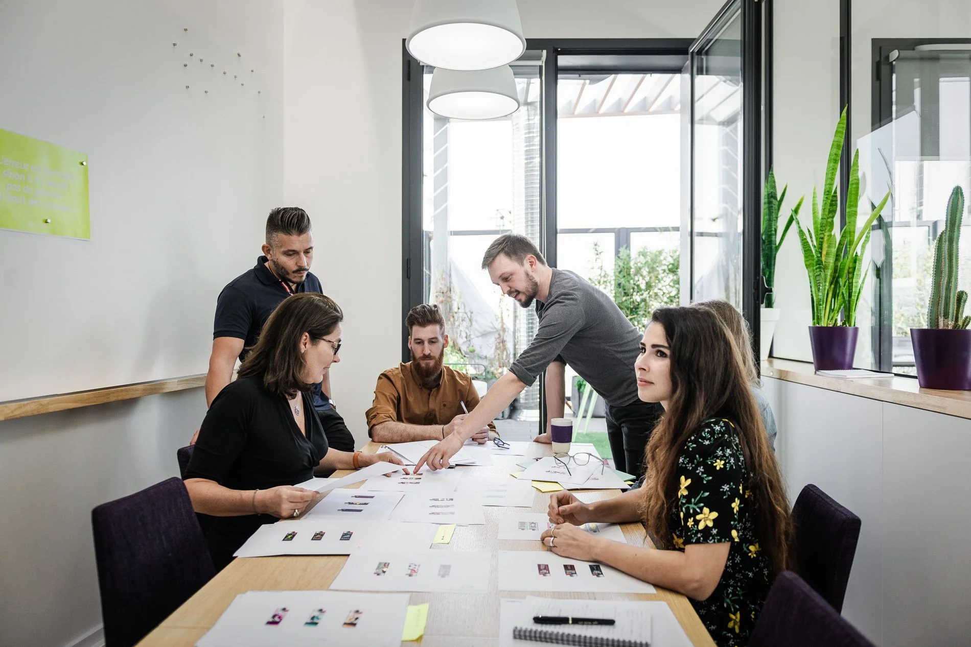 Personnes en réunion discutant autour d'une table, des documents étalés devant eux, avec des plantes en arrière-plan.